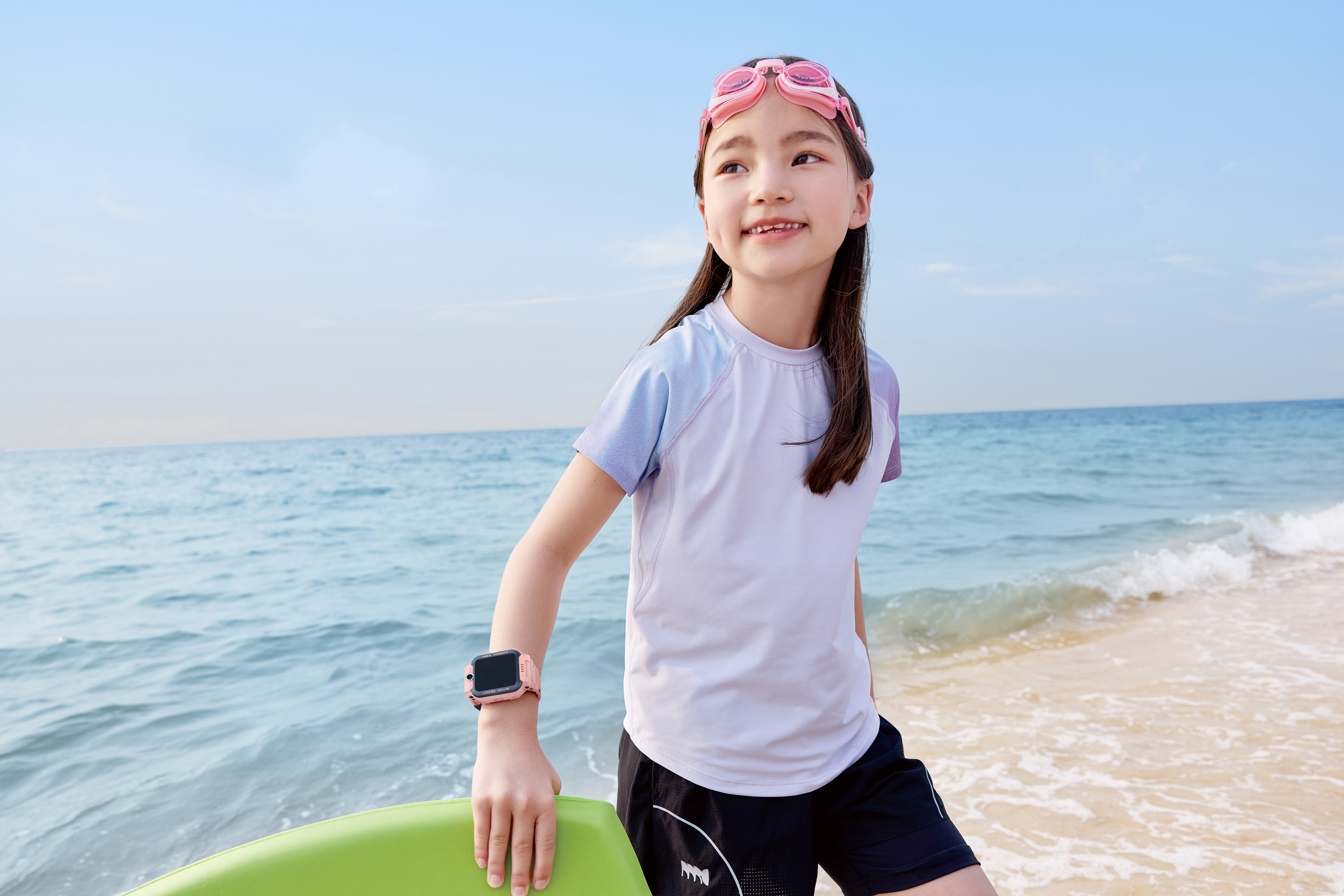 Ein Mädchen mit rosa Brille am Strand, hält ein grünes Brett, trägt eine Uhr und blickt aufs Meer.