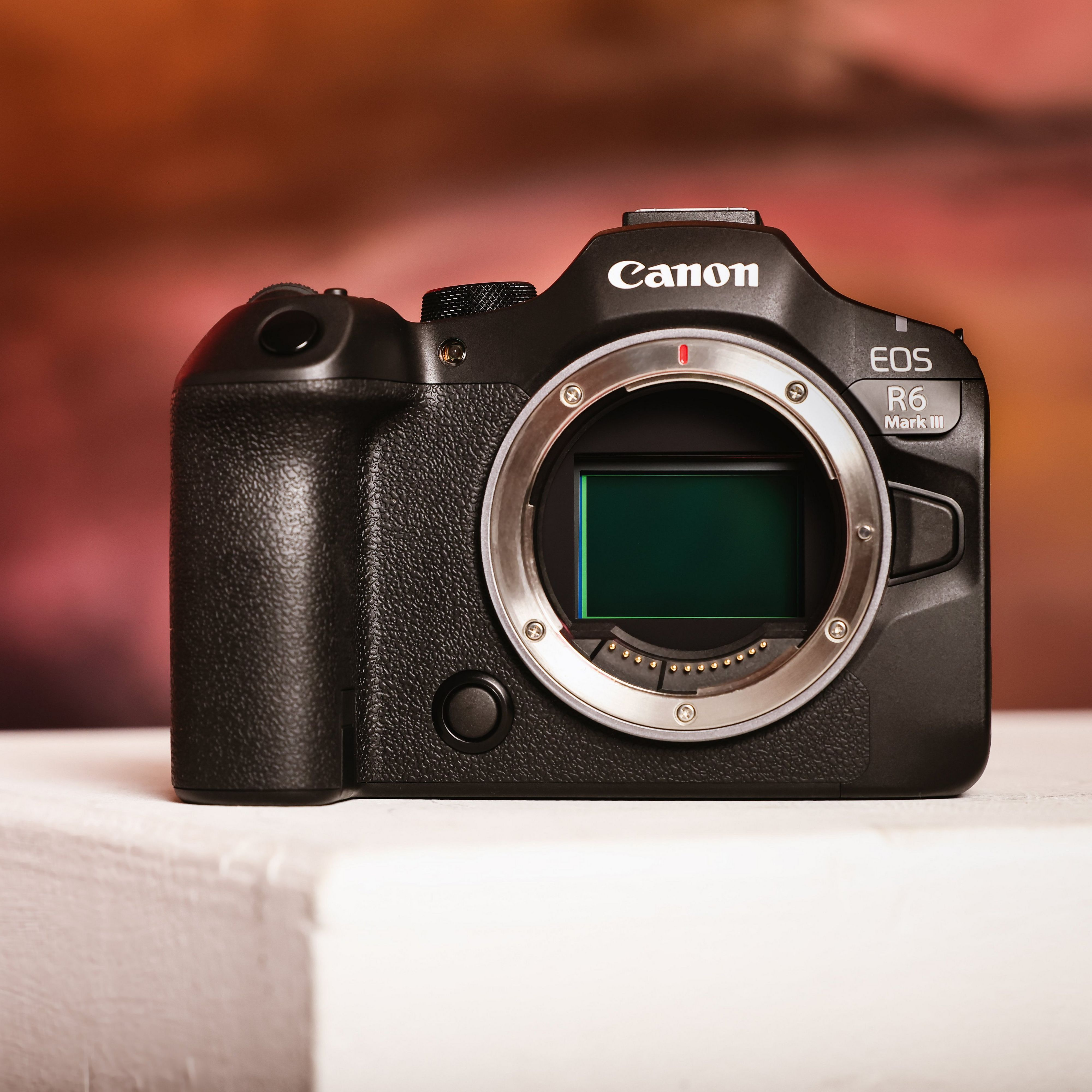 Schwarze Canon EOS R6 Mark III Kamera auf weißer Oberfläche. Der Hintergrund ist eine verschwommene Mischung aus Orange und Rot.
