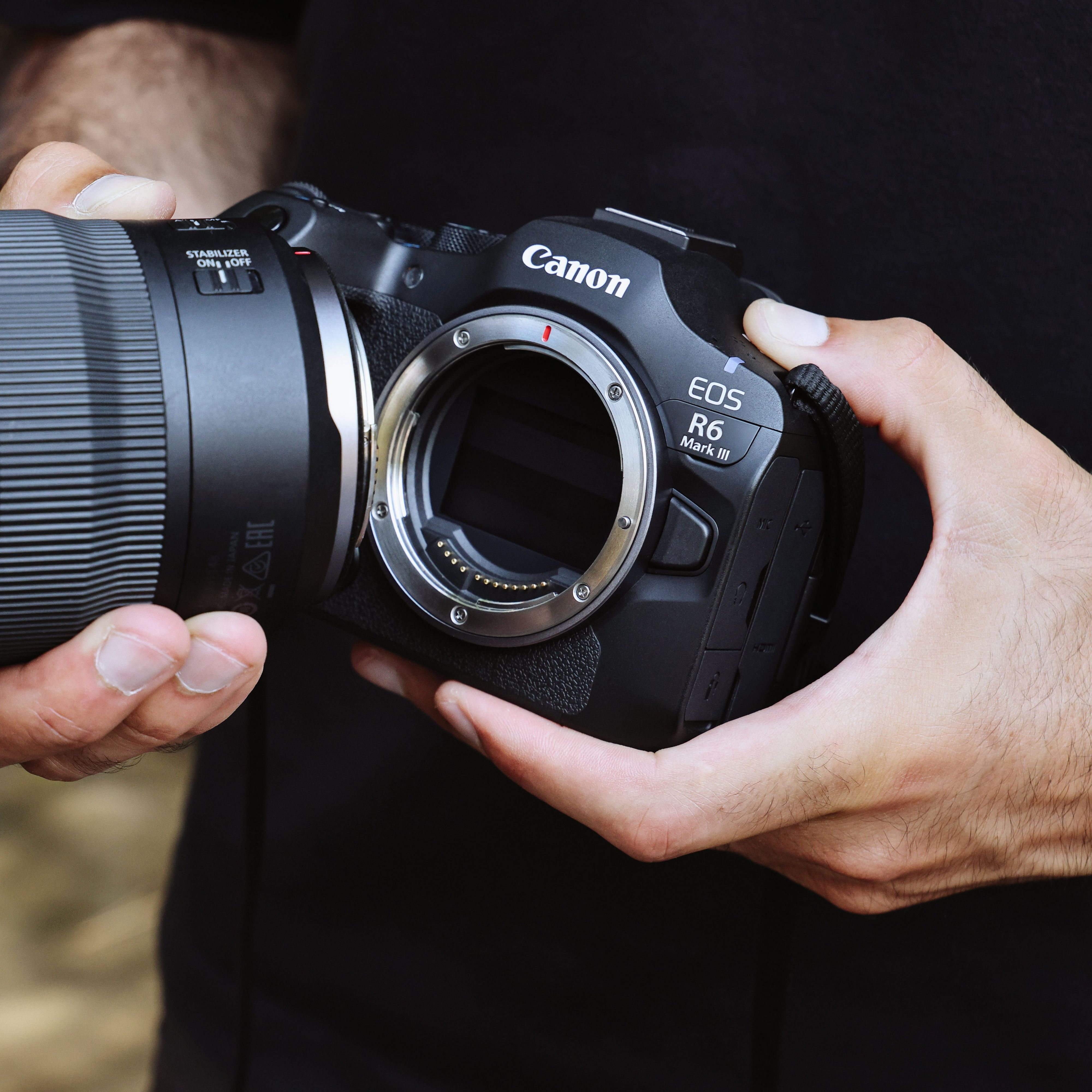 Hände halten eine Canon EOS R6 Mark III Kamera mit Objektiv. Schwarzer Hintergrund.