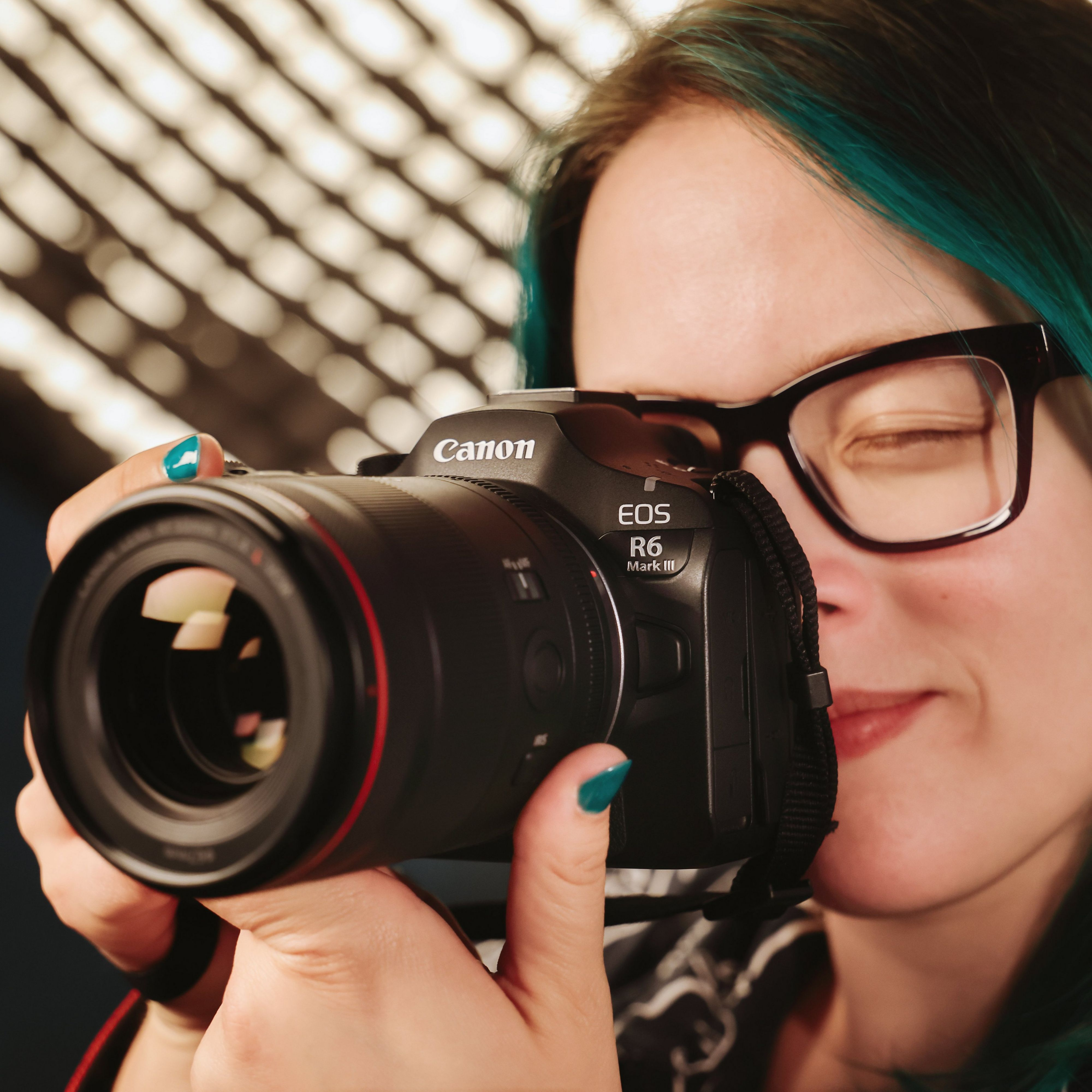 Eine Frau mit Brille und türkisfarbenem Haar hält eine Canon-Kamera. Der Hintergrund hat verschwommene Lichter. Sie lächelt.