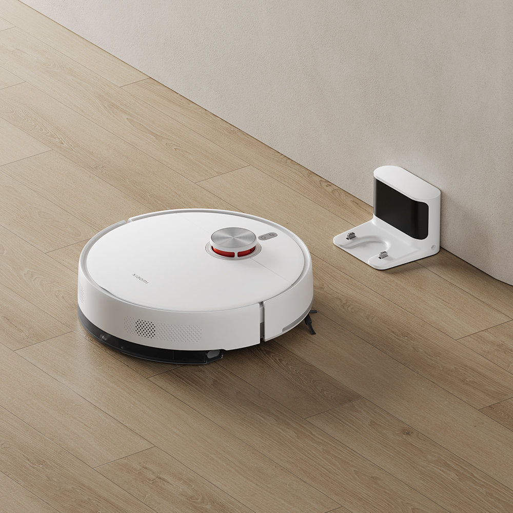 Un robot aspirapolvere bianco e la sua base di ricarica su un pavimento in legno, vicino a un muro. Il robot ha un pulsante rosso.