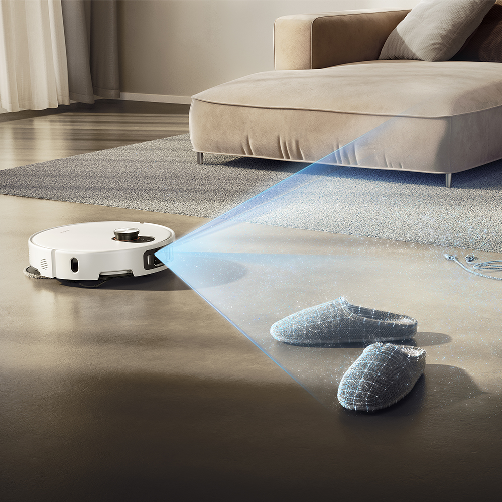 Un aspirateur robot blanc nettoie un salon. Des pantoufles grises sont sur le sol. Un canapé et un tapis sont en arrière-plan.