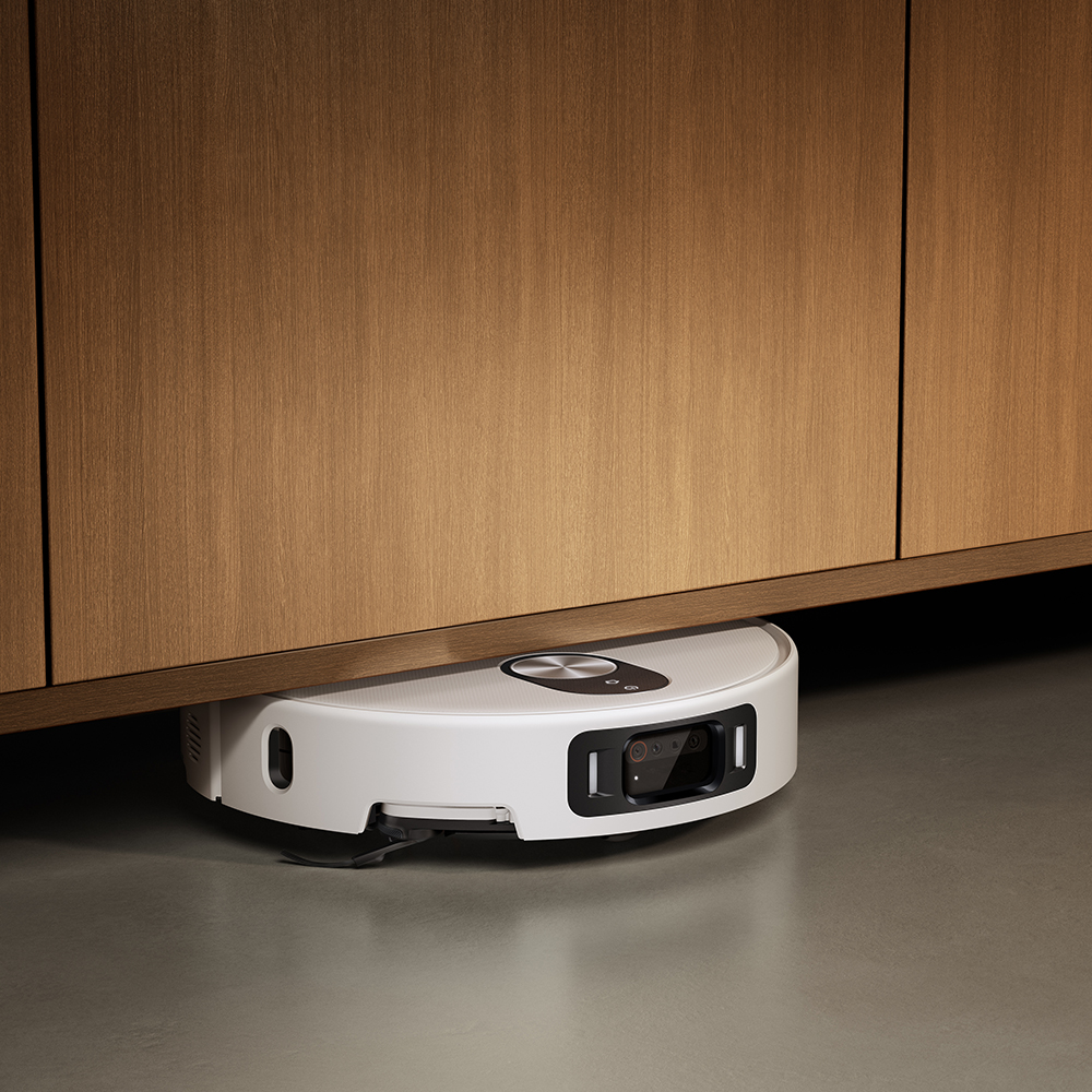 Un aspirateur robot blanc nettoie sous une armoire en bois. Il est sur un sol gris. L'armoire est marron.