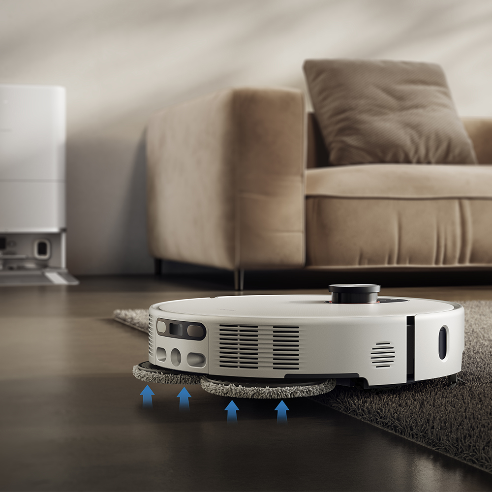 Un aspirateur robot blanc nettoie un tapis. Des flèches indiquent sa direction de nettoyage. Un canapé et un appareil blanc en arrière-plan.