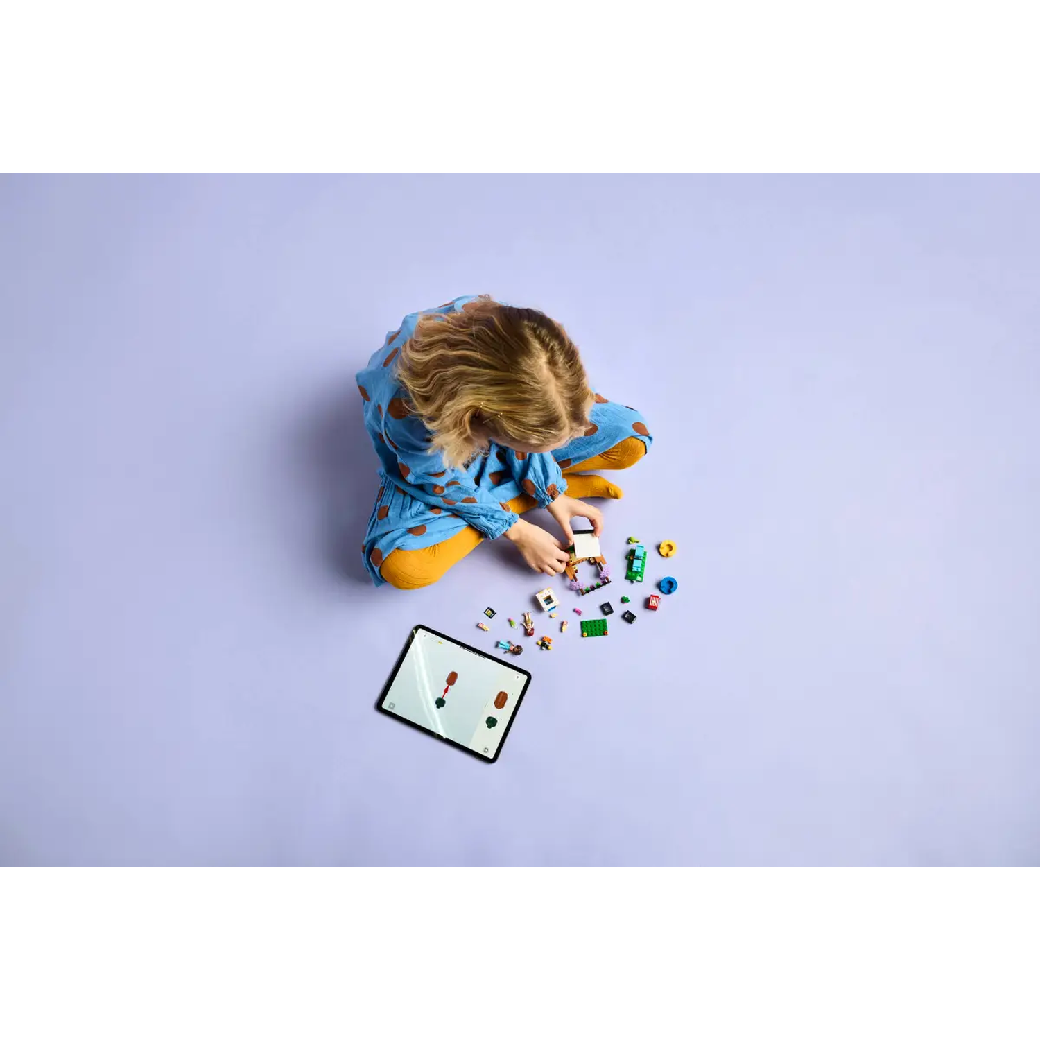 A child sits on purple ground playing with blocks and a tablet. The child wears blue with brown spots and yellow pants.