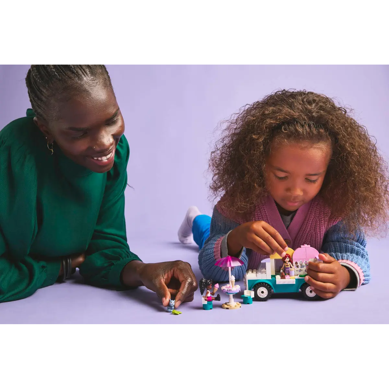 Een vrouw en een meisje spelen met LEGO Friends-speelgoed op een paars oppervlak. De set bevat een foodtruck en kleine figuren.