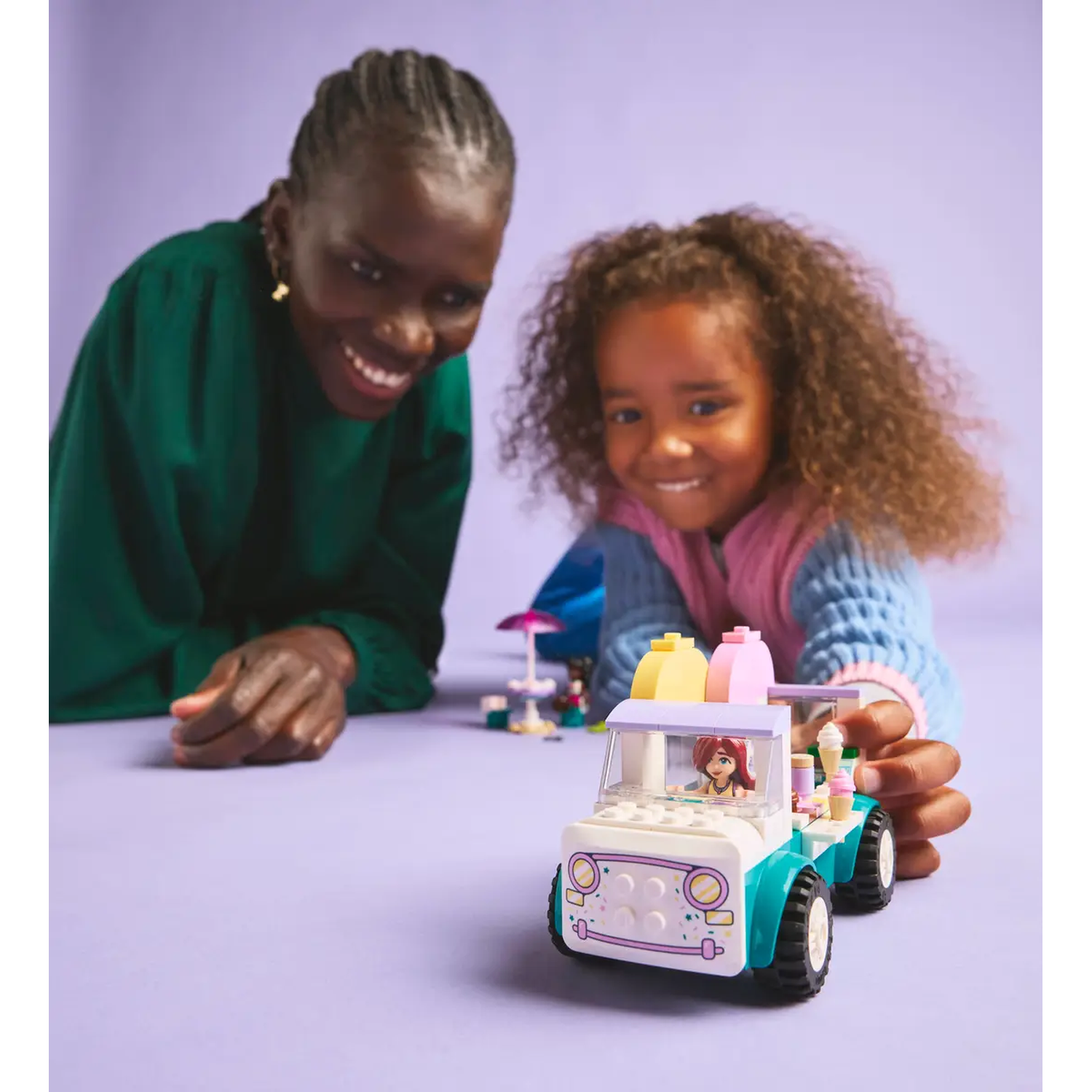 Een vrouw en een meisje spelen met een Lego-ijscowagen. De wagen heeft ijs bovenop. Beiden lachen naar de camera.