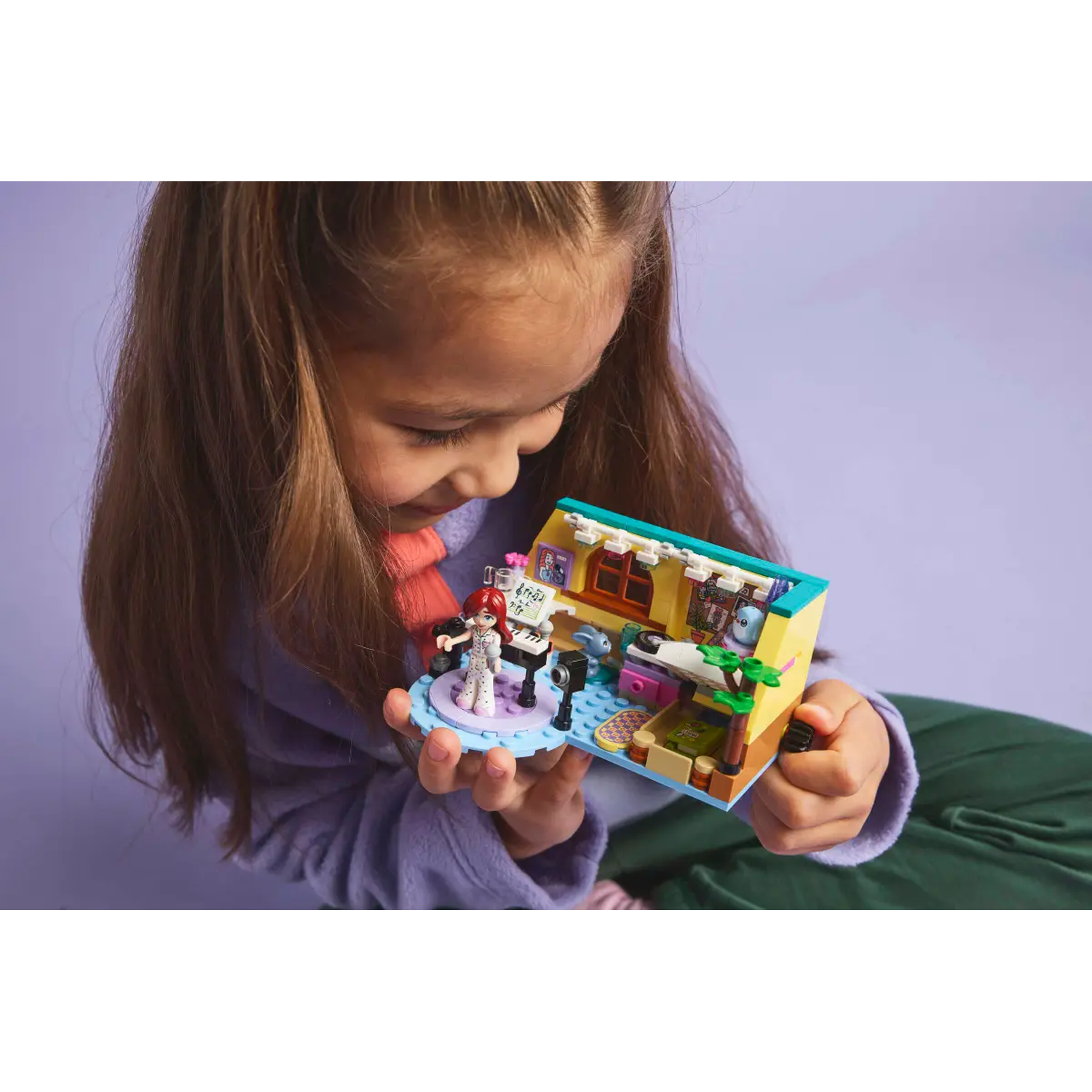 A girl holds a Lego set. The set shows a stage with a singer and a small house with a bedroom. She looks at it closely.