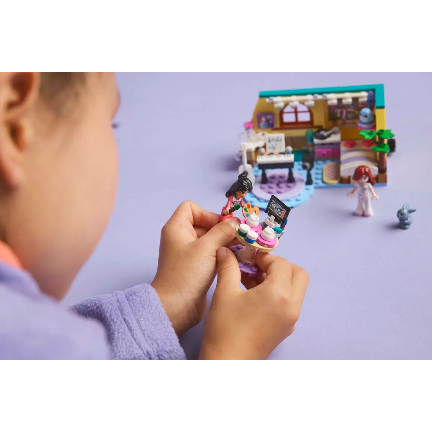A child holds a LEGO set with figures and a building. The background shows the house and other figures on a purple surface.