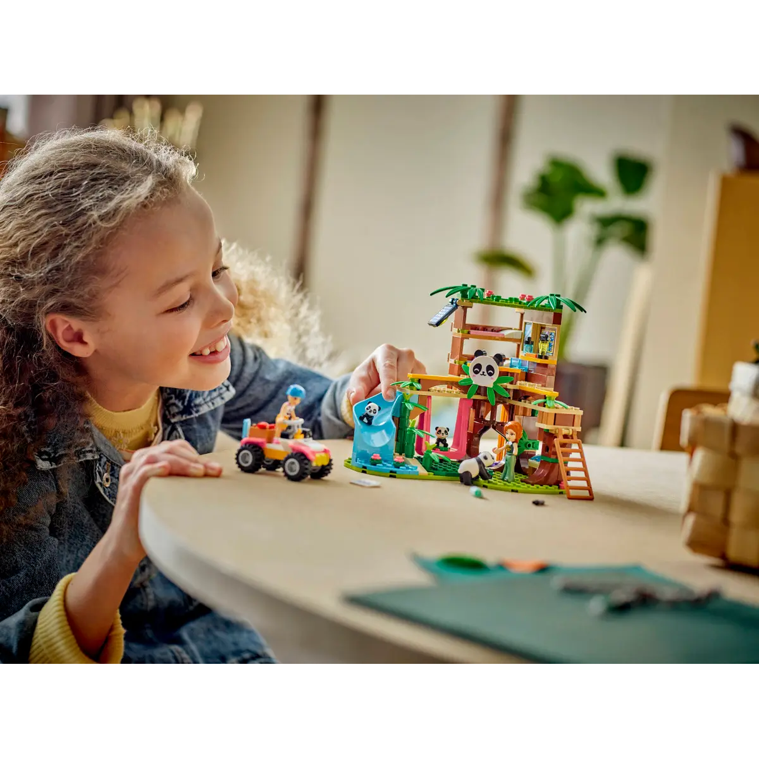 Een meisje speelt met een Lego-set met panda's en een boomhut. Er staat een kleine auto op tafel. Het meisje lacht.
