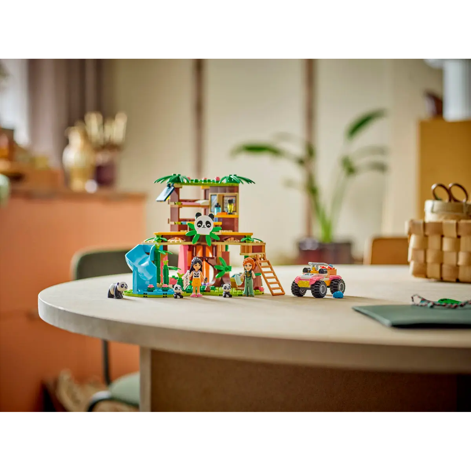 Een Lego-set met een panda-boomhut, figuren, panda's en een auto, op een houten tafel. Een plant en een mand op de achtergrond.