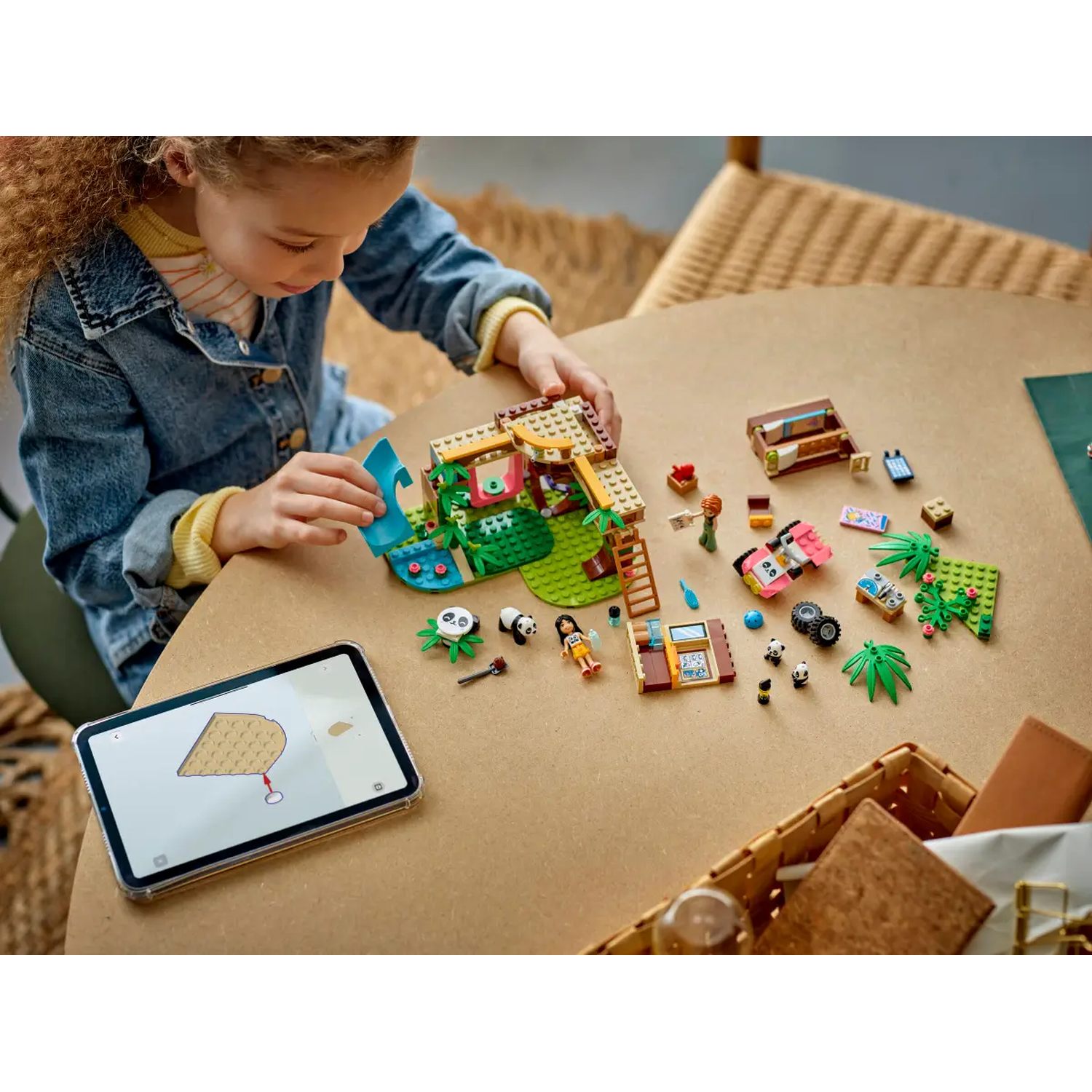 Een meisje bouwt een Lego-set op een tafel. De set bevat een huis, een auto, panda's en andere kleine items. Een tablet toont instructies.