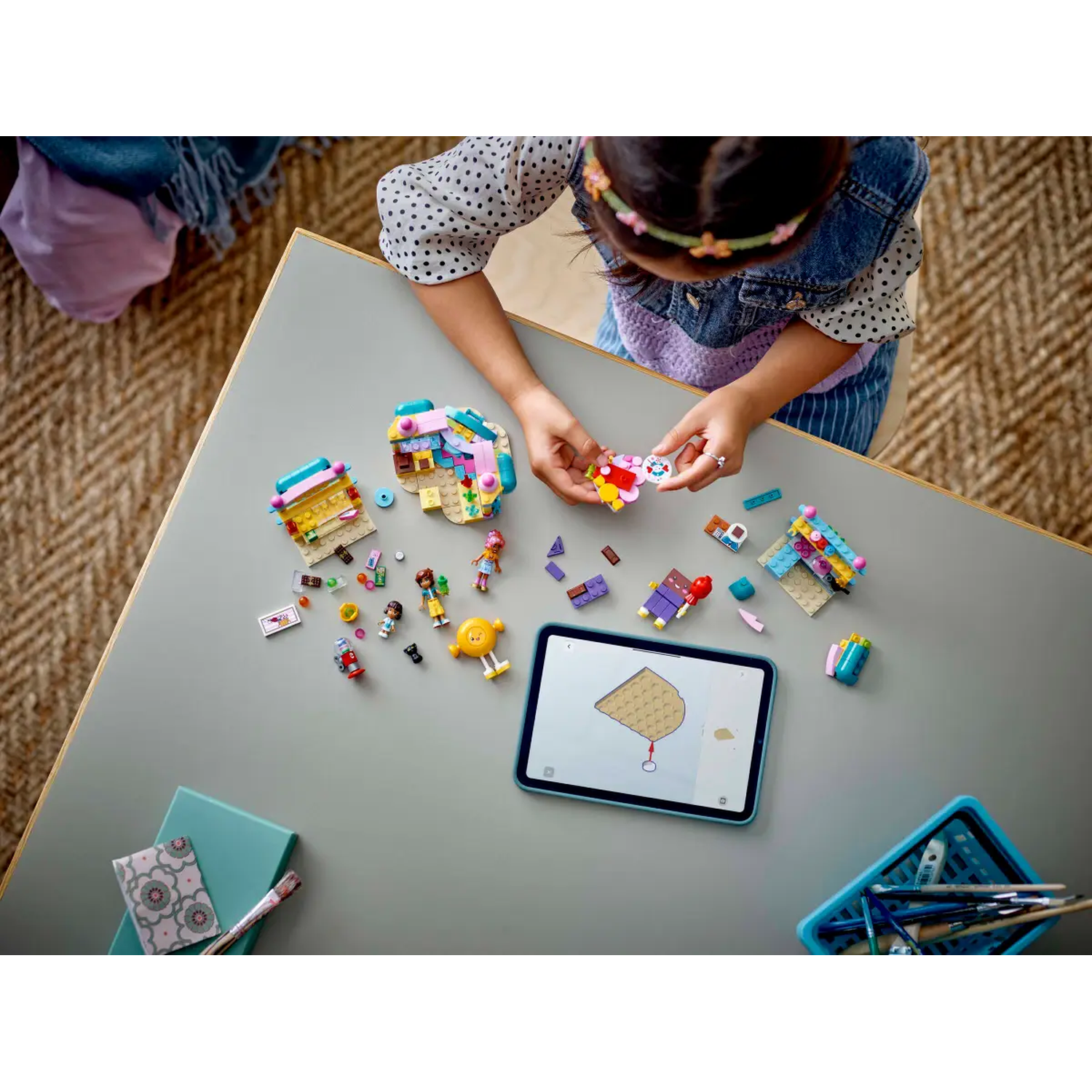 A child is building with colorful Lego bricks on a gray table. There is a tablet showing instructions and other toys.