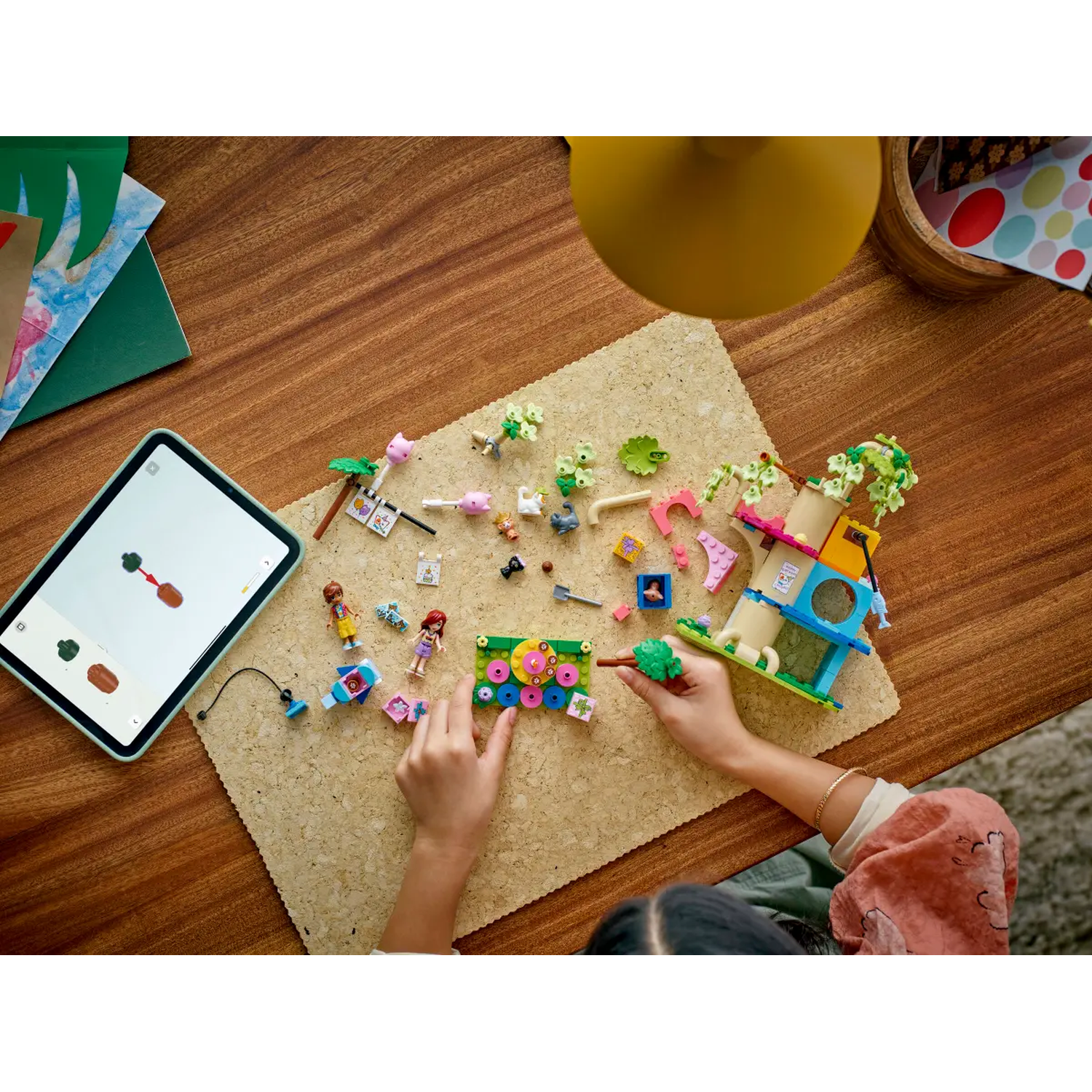 A person builds a colorful Lego set on a corkboard. A tablet shows instructions. Various Lego pieces are scattered around the board.