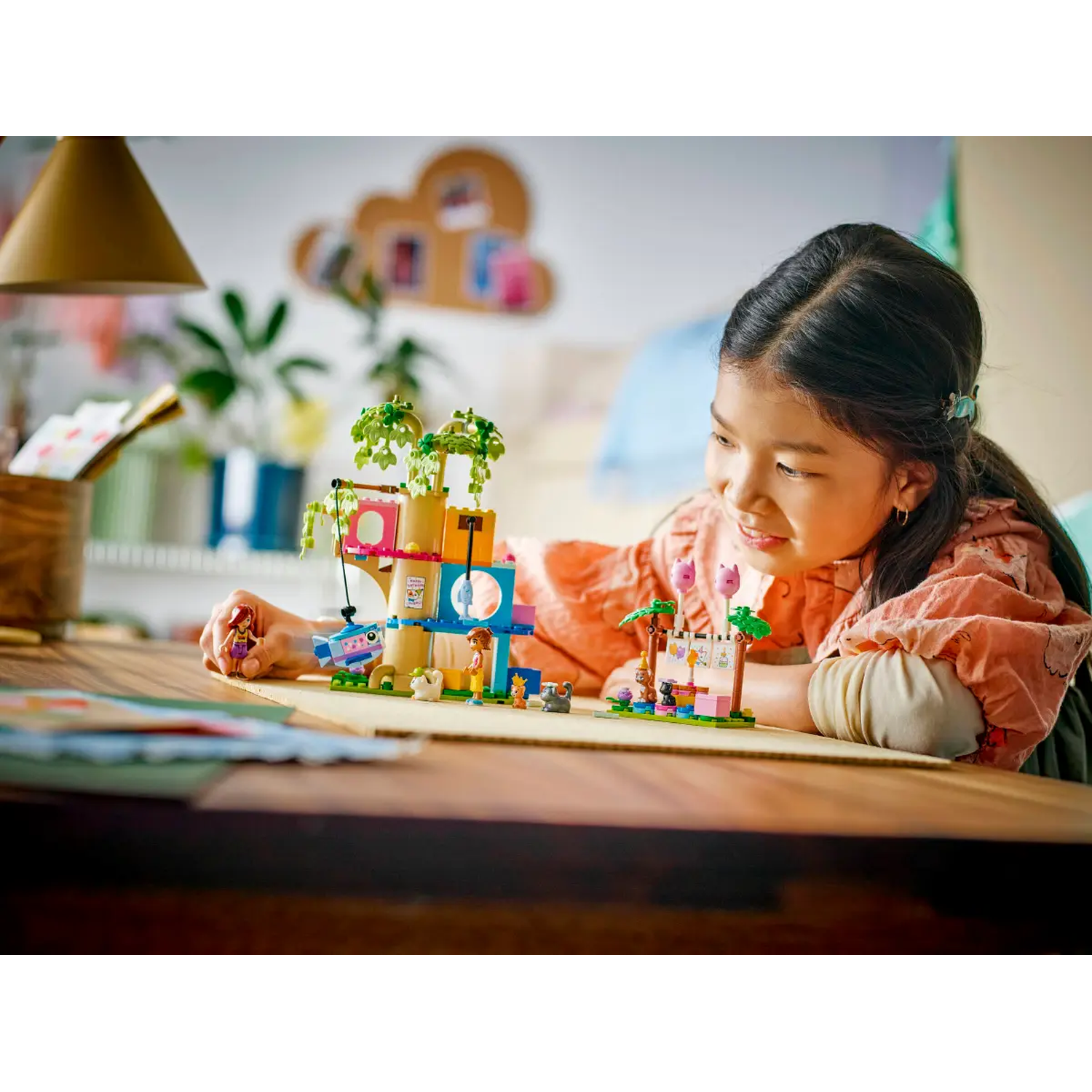 A girl looks at a Lego set on a desk. The set includes a treehouse and other small Lego figures and constructions.