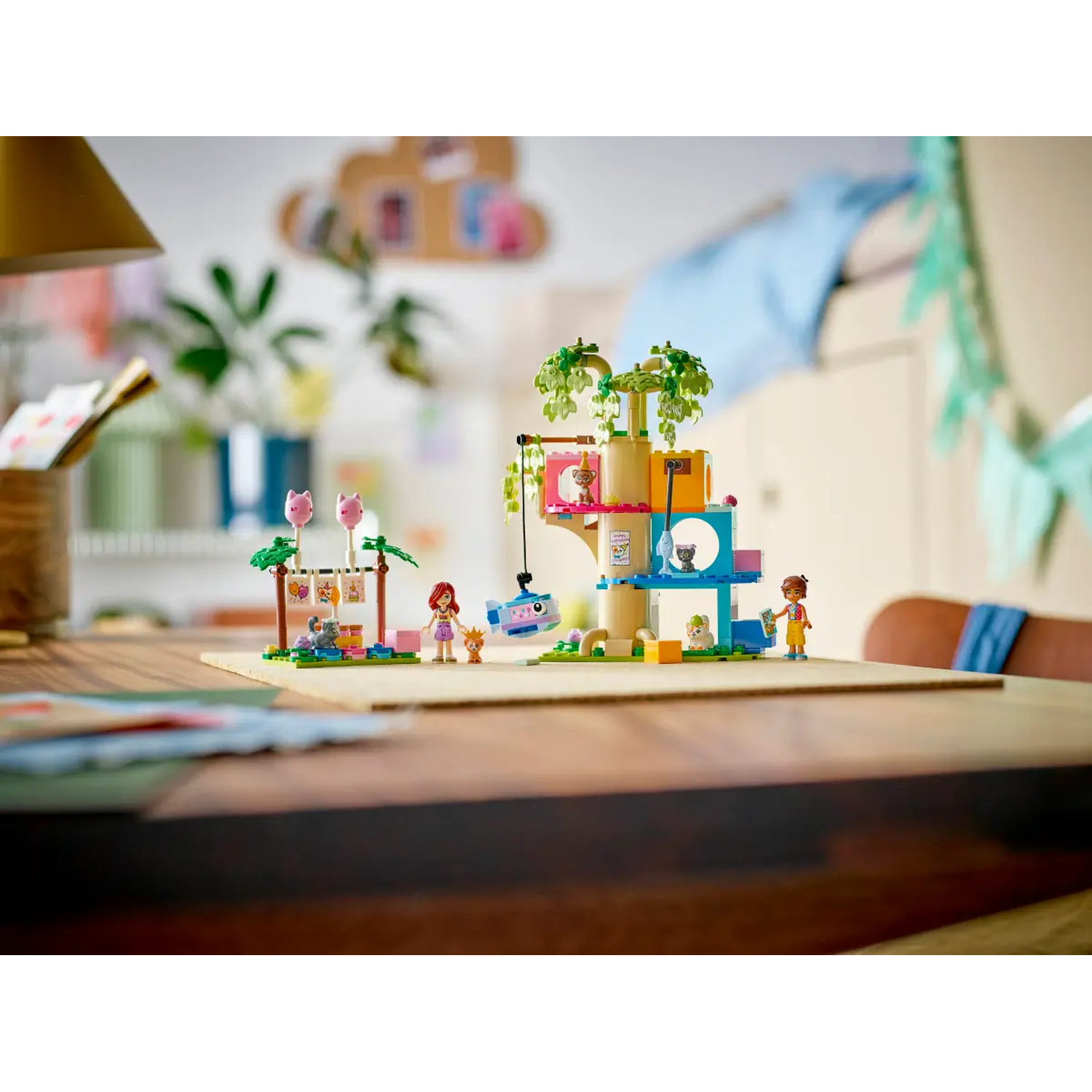 A Lego cat tree with figures. Cat-shaped balloons and a banner in the background. A wooden table with paper in the foreground.