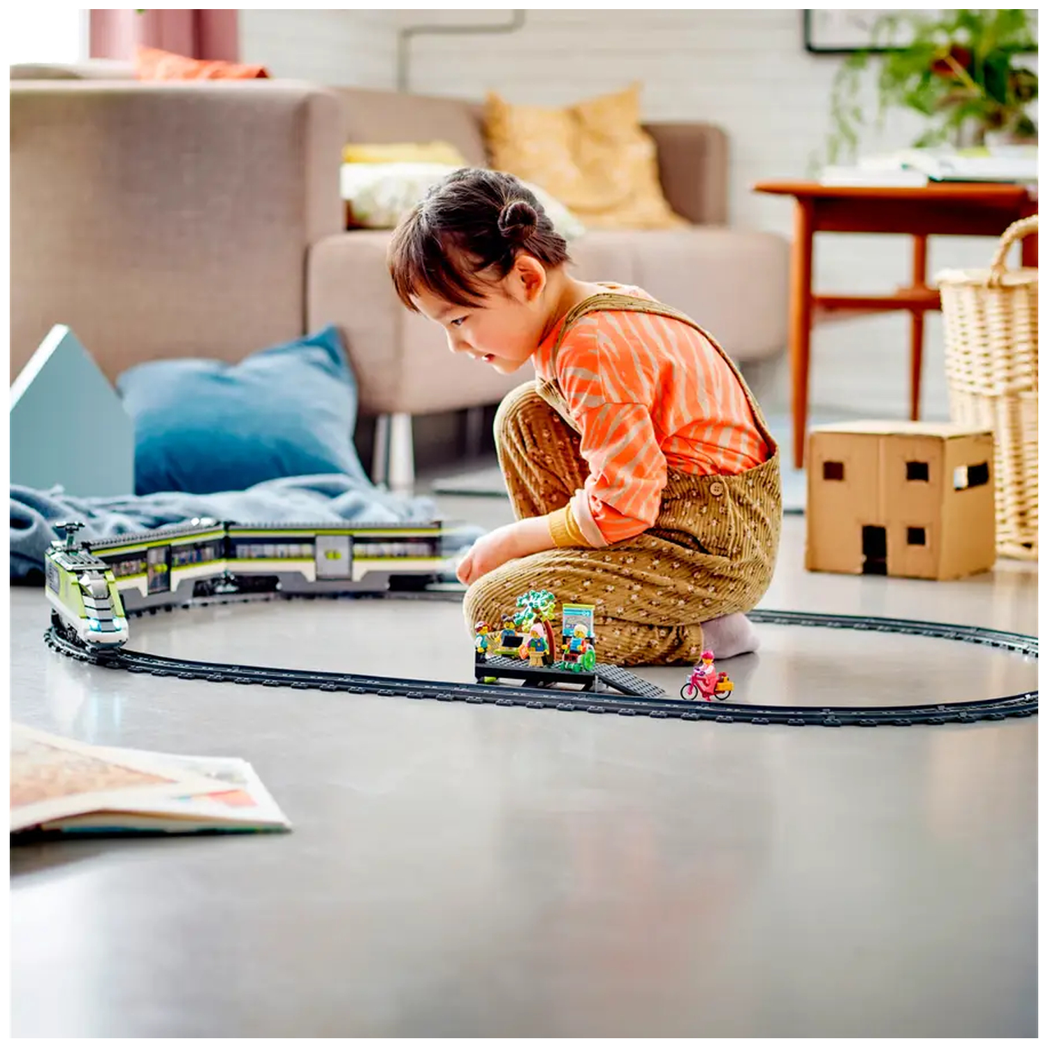 Een kind speelt met een speelgoedtreinset, met een baan op de grond. Het kind draagt een overall. Op de achtergrond een bank en andere speeltjes.