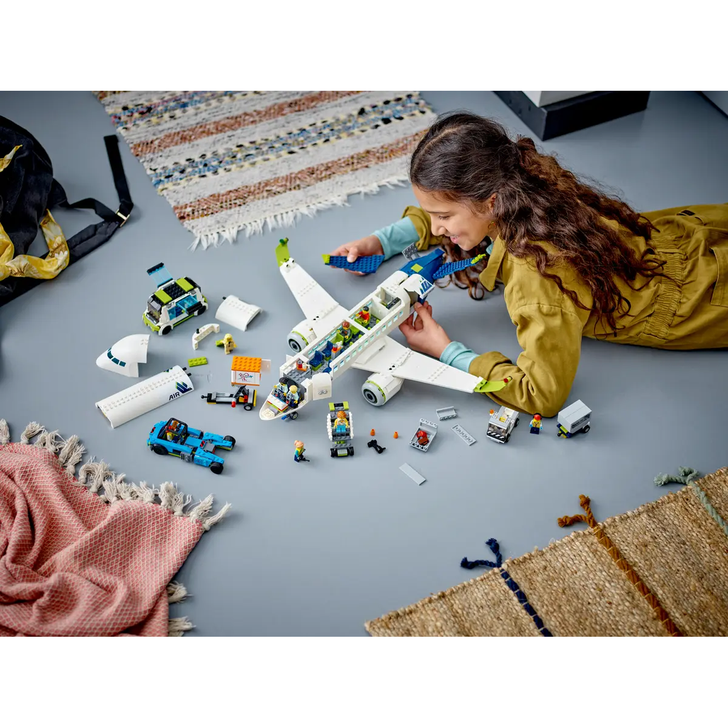A girl plays with a Lego airplane set. The set includes a plane, a truck, and other airport-related elements. The girl is smiling.