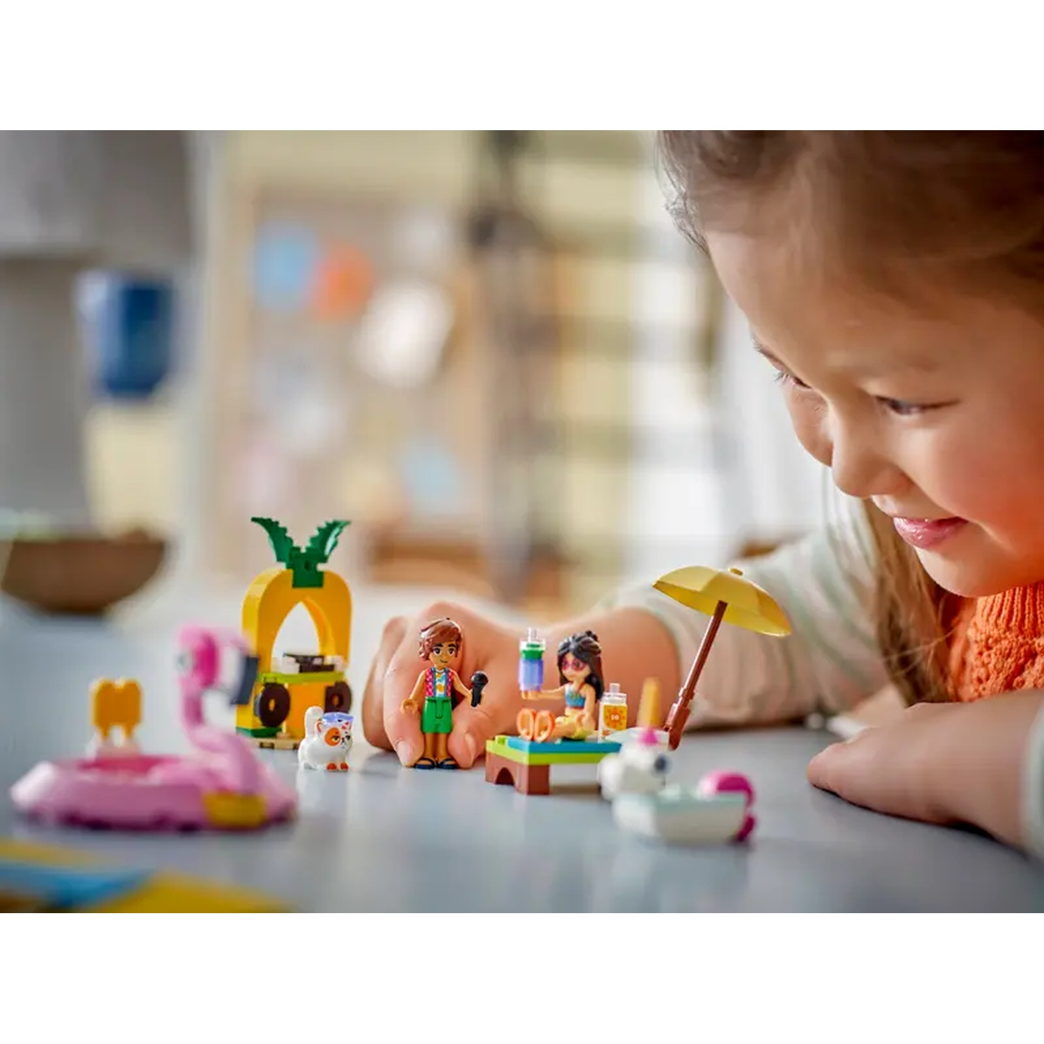 A young girl looks at a Lego set featuring a beach scene with figurines, a pineapple stand, and a flamingo float.