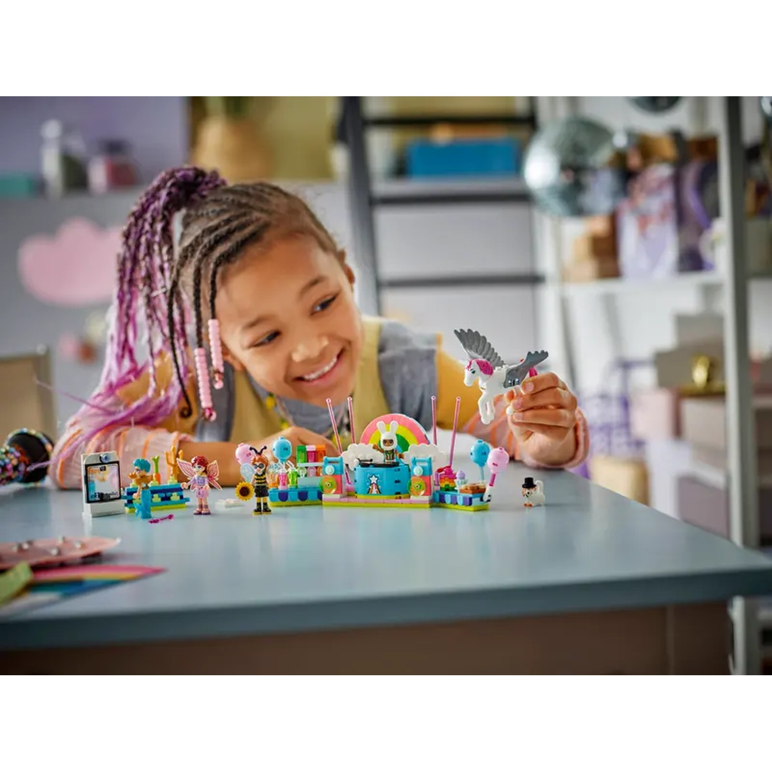 A girl smiles, holding a toy unicorn. A colorful Lego set is on the table, with various figures and structures. Background includes shelves.