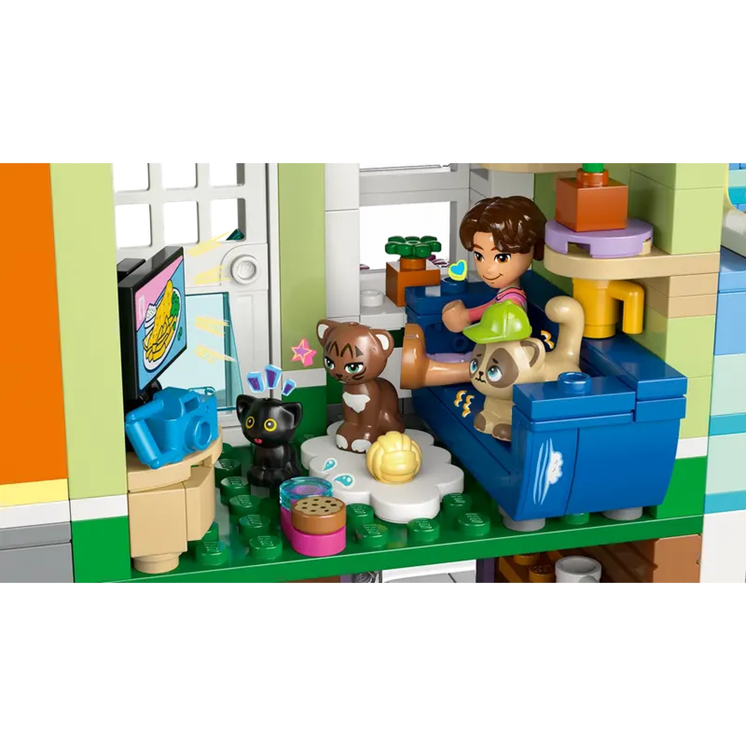 Een Lego-scène met een persoon op een blauwe bank met twee kattenfiguren. Er zijn andere kattenfiguren en objecten. De tv toont eten.