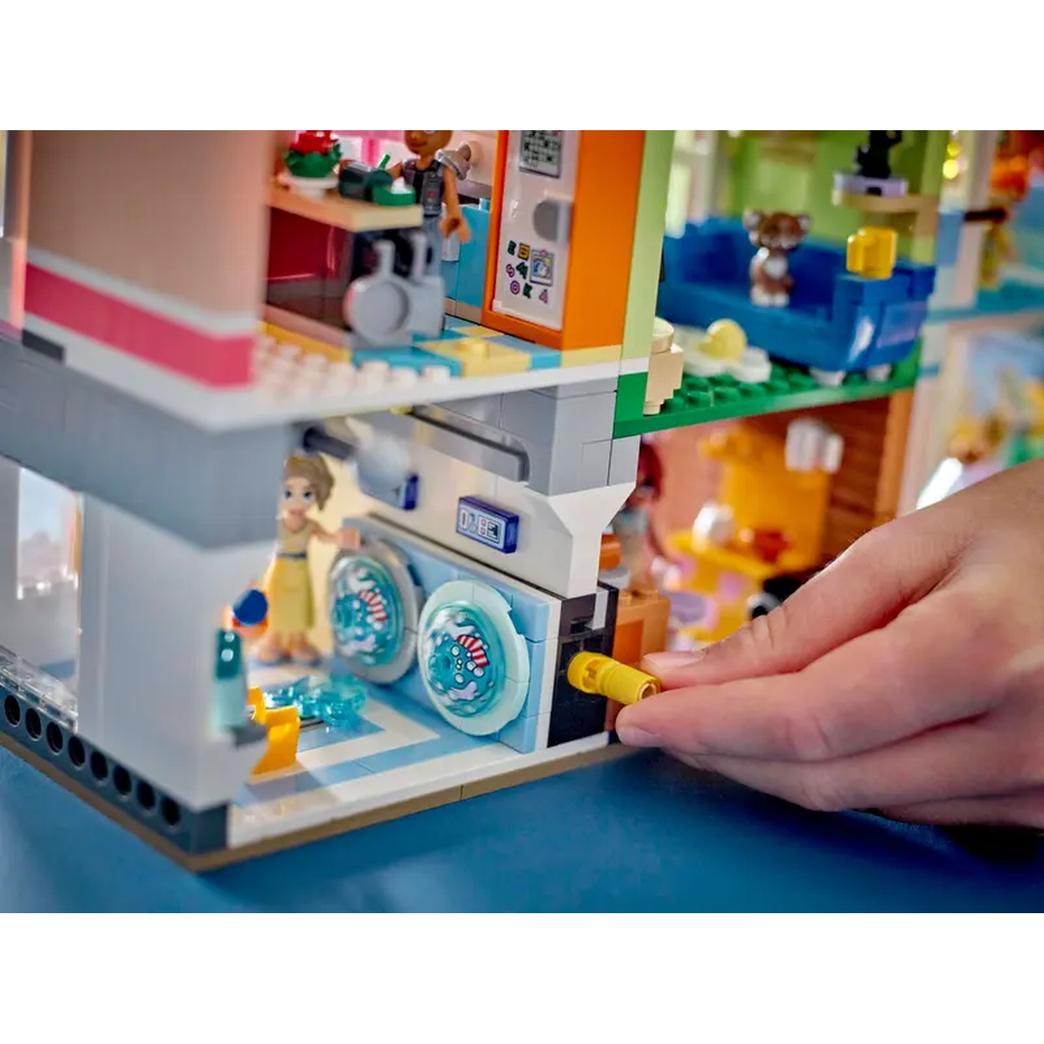Lego-set met een gebouw met twee verdiepingen. Een hand houdt een geel stuk vast. Figuren zijn aanwezig in verschillende kamers met meubels.