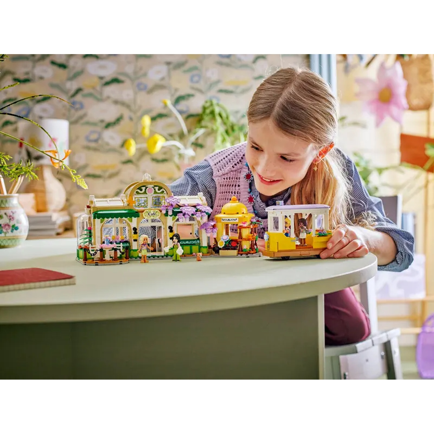 A girl smiles at a Lego set on a table. The set includes buildings and figures. Floral wallpaper is in the background.