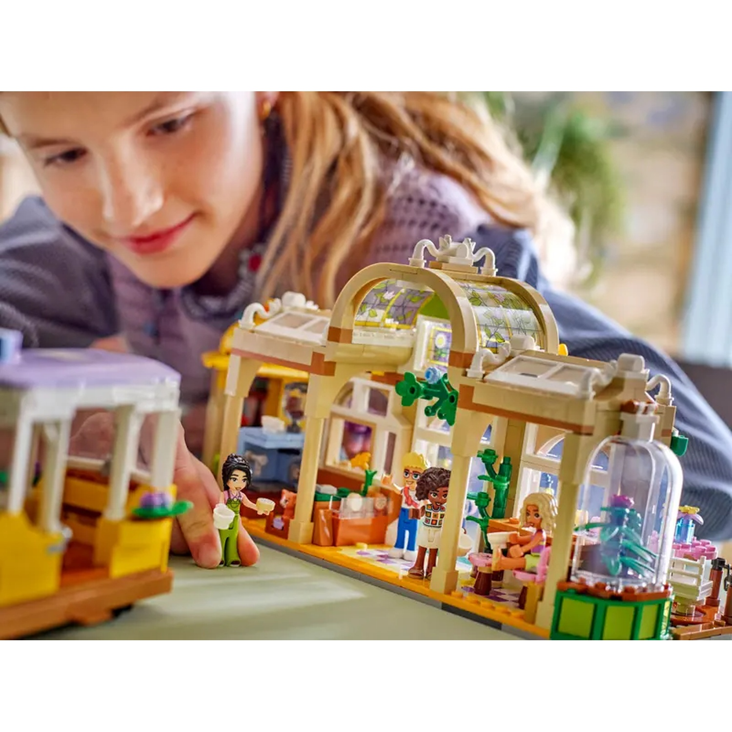 A girl looks at a Lego set with a greenhouse, figures, and a yellow and purple trolley. The set is colorful.