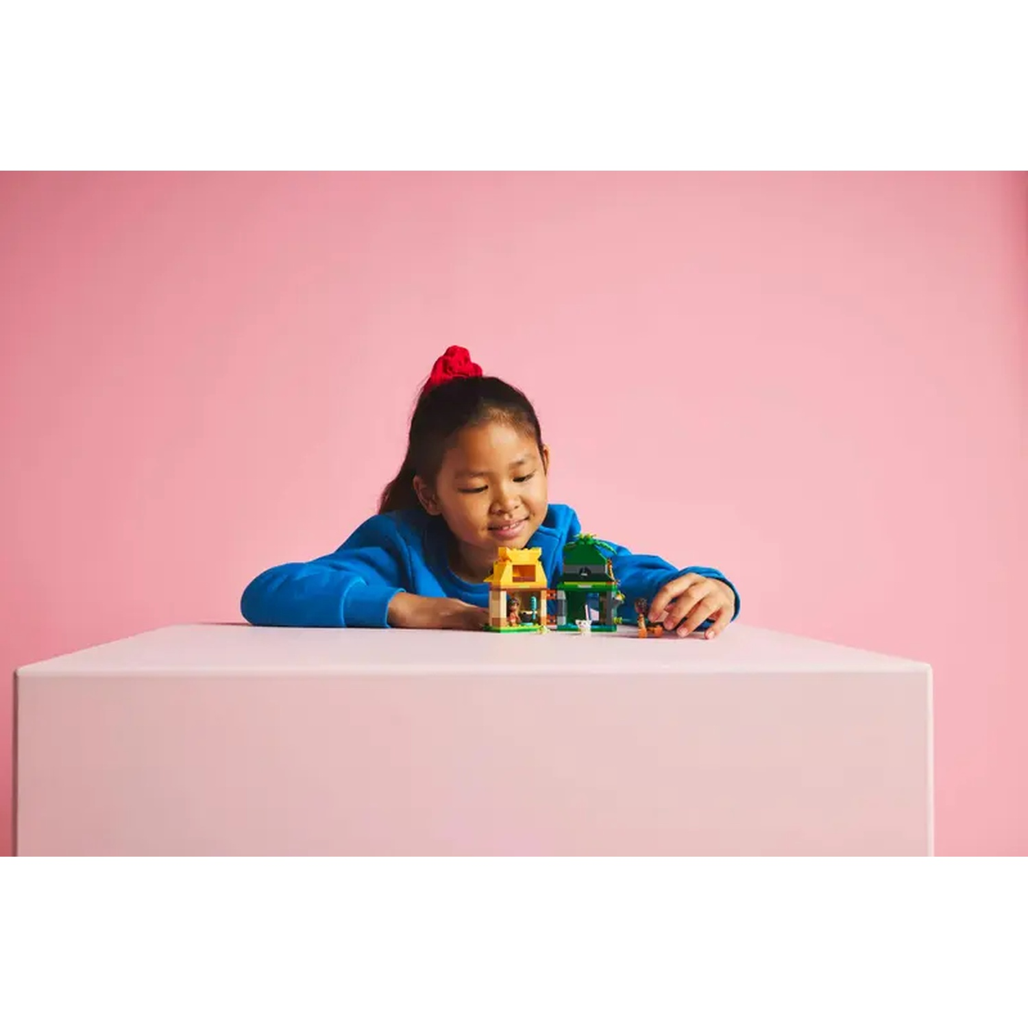 Une fille avec un nœud rouge regarde des structures Lego sur une table rose. Le fond est rose.