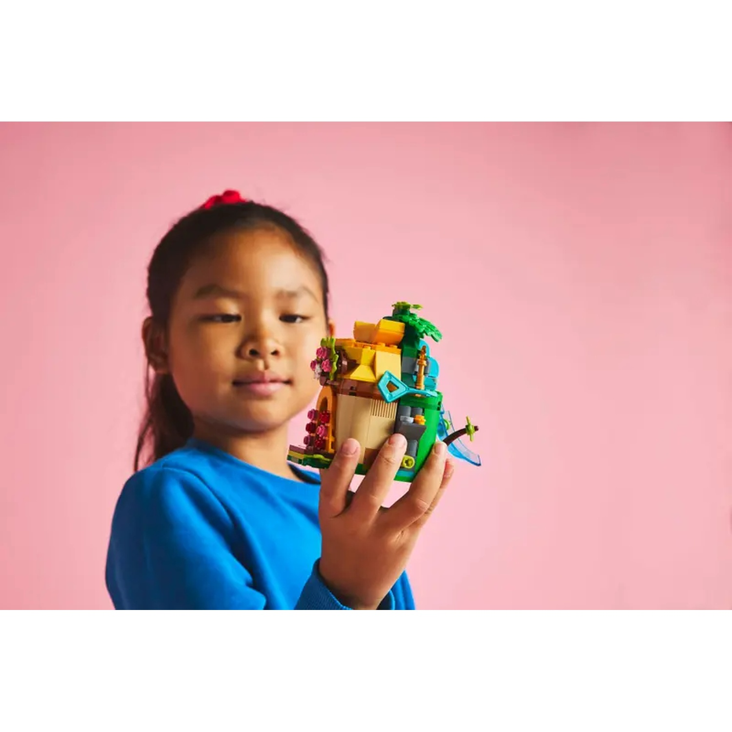 Une jeune fille en bleu tient une création Lego avec des couleurs jaune, vert et marron, sur fond rose.