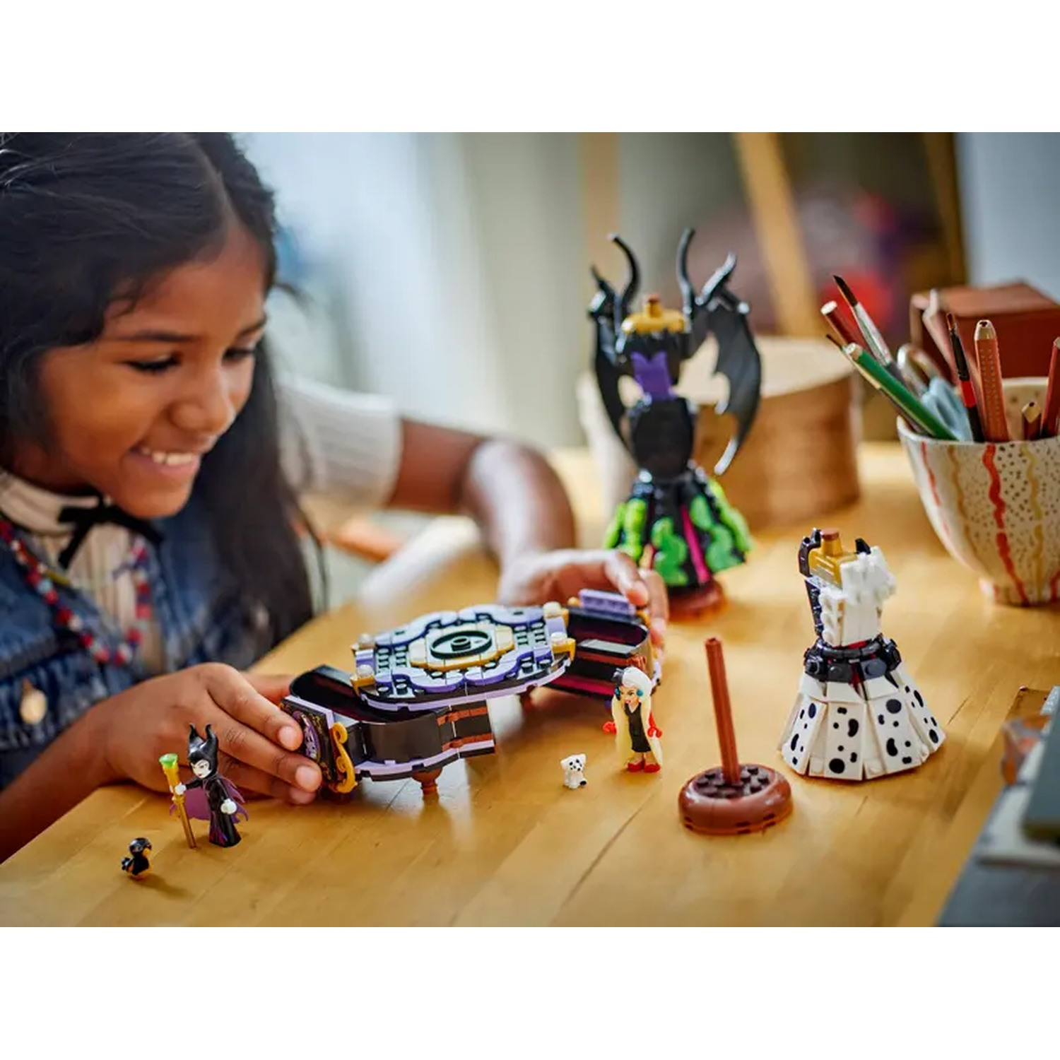 Een meisje glimlacht naar Lego-figuren op een houten tafel. Lego-figuren zijn onder meer Maleficent, Cruella en een Dalmatische jurk.