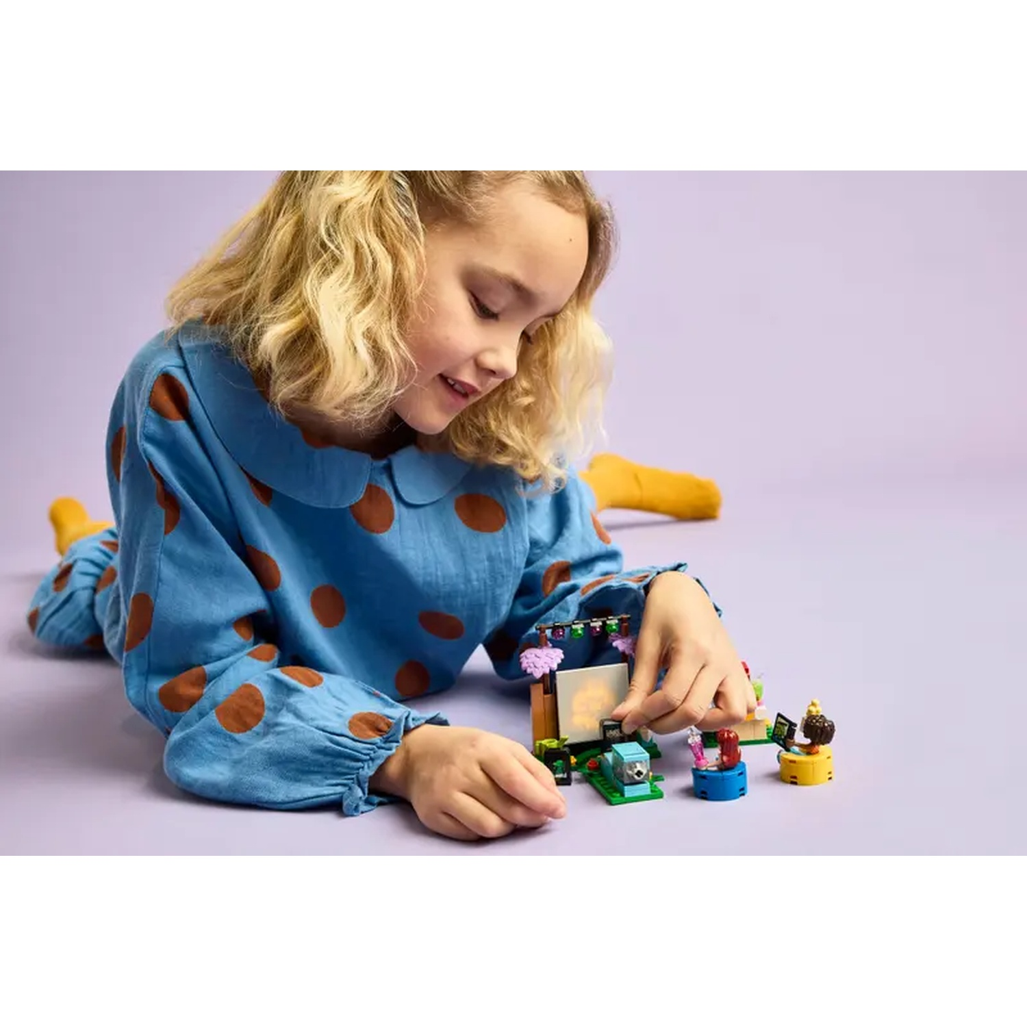 A girl in blue polka-dot pajamas plays with Lego blocks. Various Lego figures and a screen are visible.