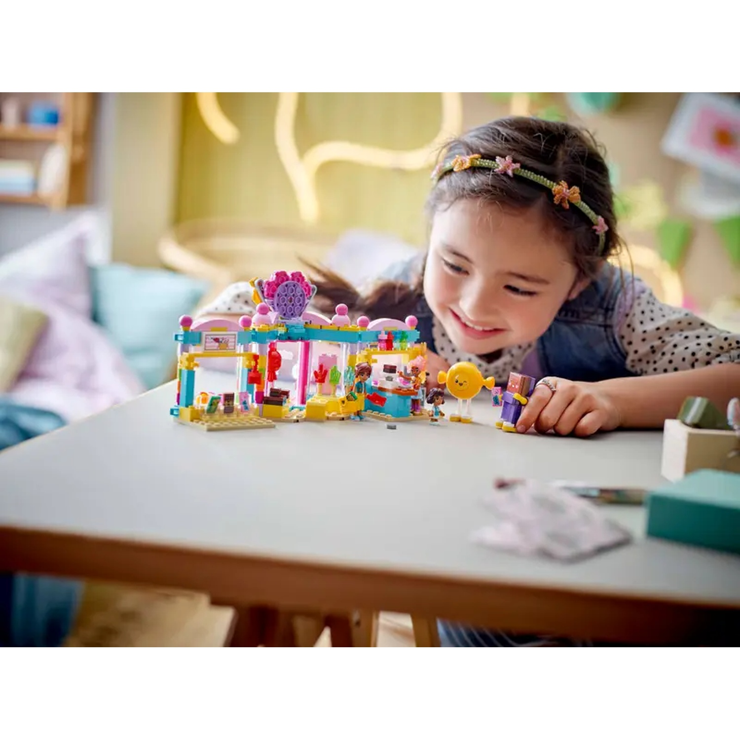 A girl smiles at a Lego set on a table. The set is a colorful shop. A yellow figure is near the girl's hand.