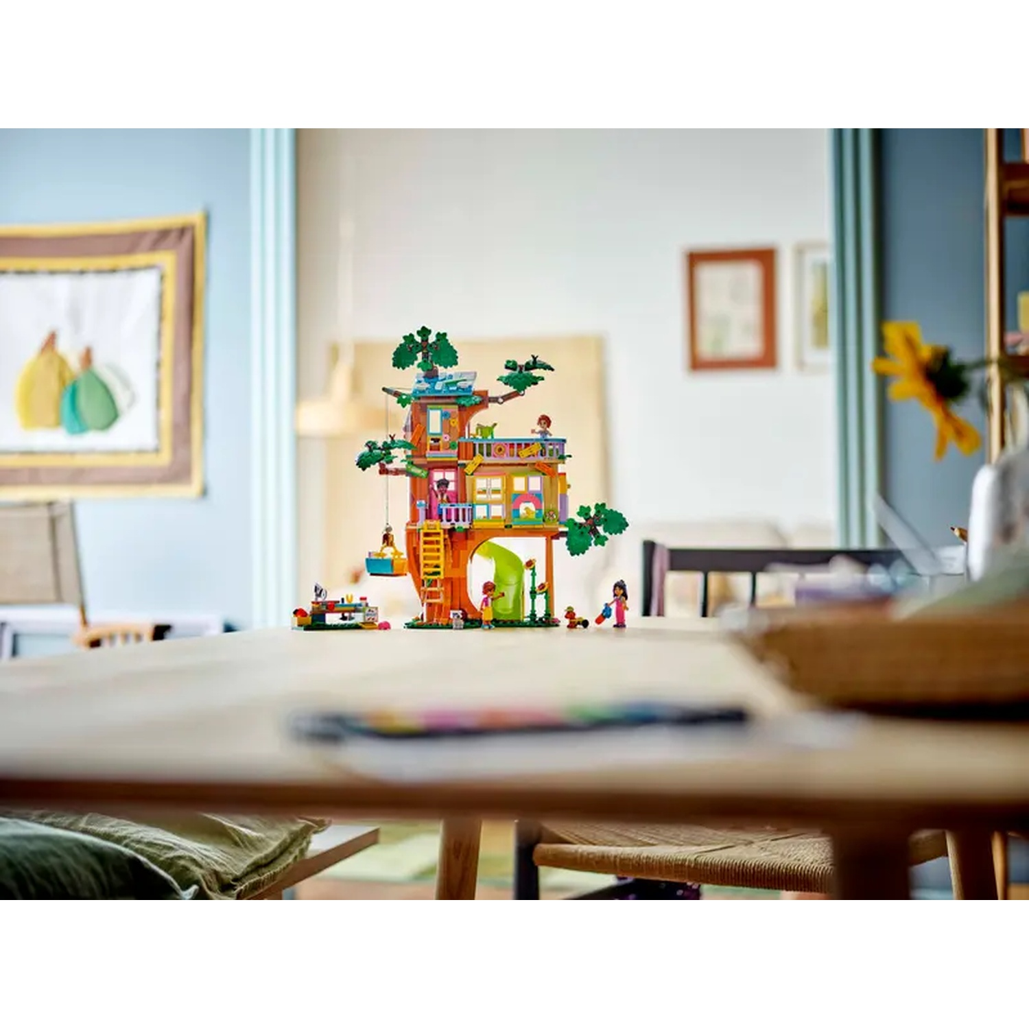 Une cabane dans les arbres Lego avec des figurines est sur une table en bois. Une image est accrochée au mur et une autre pièce est visible en arrière-plan.