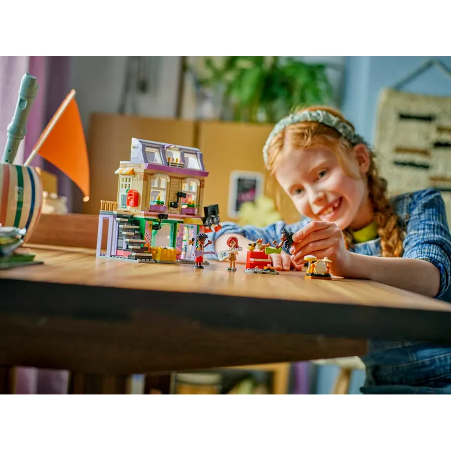 Een meisje glimlacht naar een Lego-set. Een gebouw en beeldjes staan op de tafel. Links is een oranje vlag.
