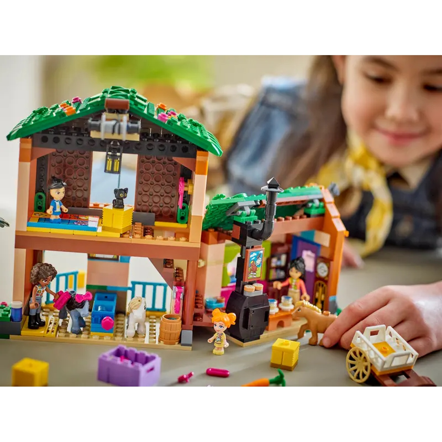 Lego-set met een huis met twee verdiepingen, figuren, dieren en een kar. Een meisje speelt met het speelgoed.