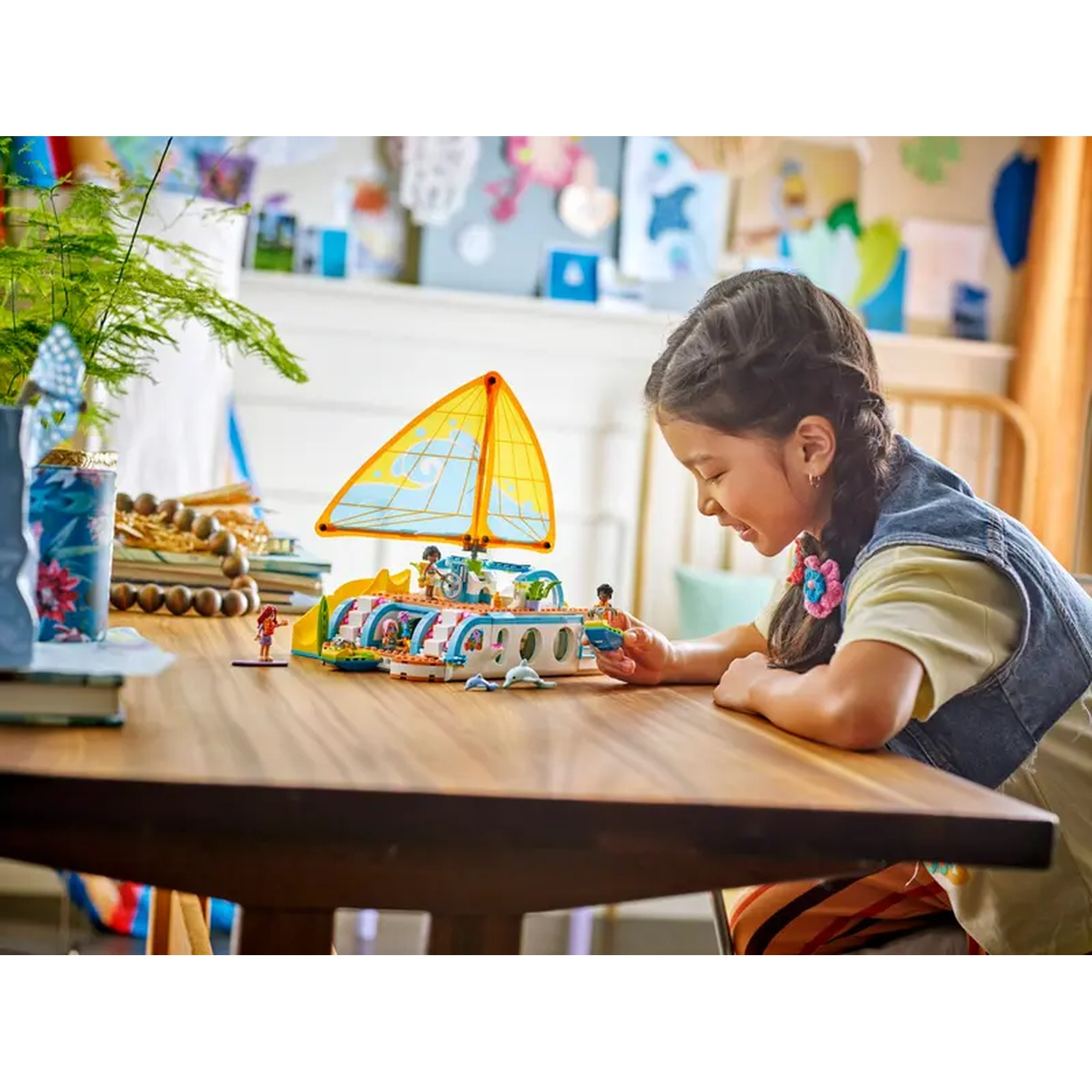 A girl looks at a Lego boat on a table. The boat has a yellow sail. Plants and drawings are in the background.