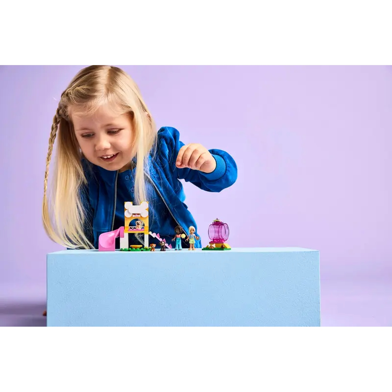 A young girl looks at a Lego set with toy figures and a dog house. The background is a light purple with a blue box.