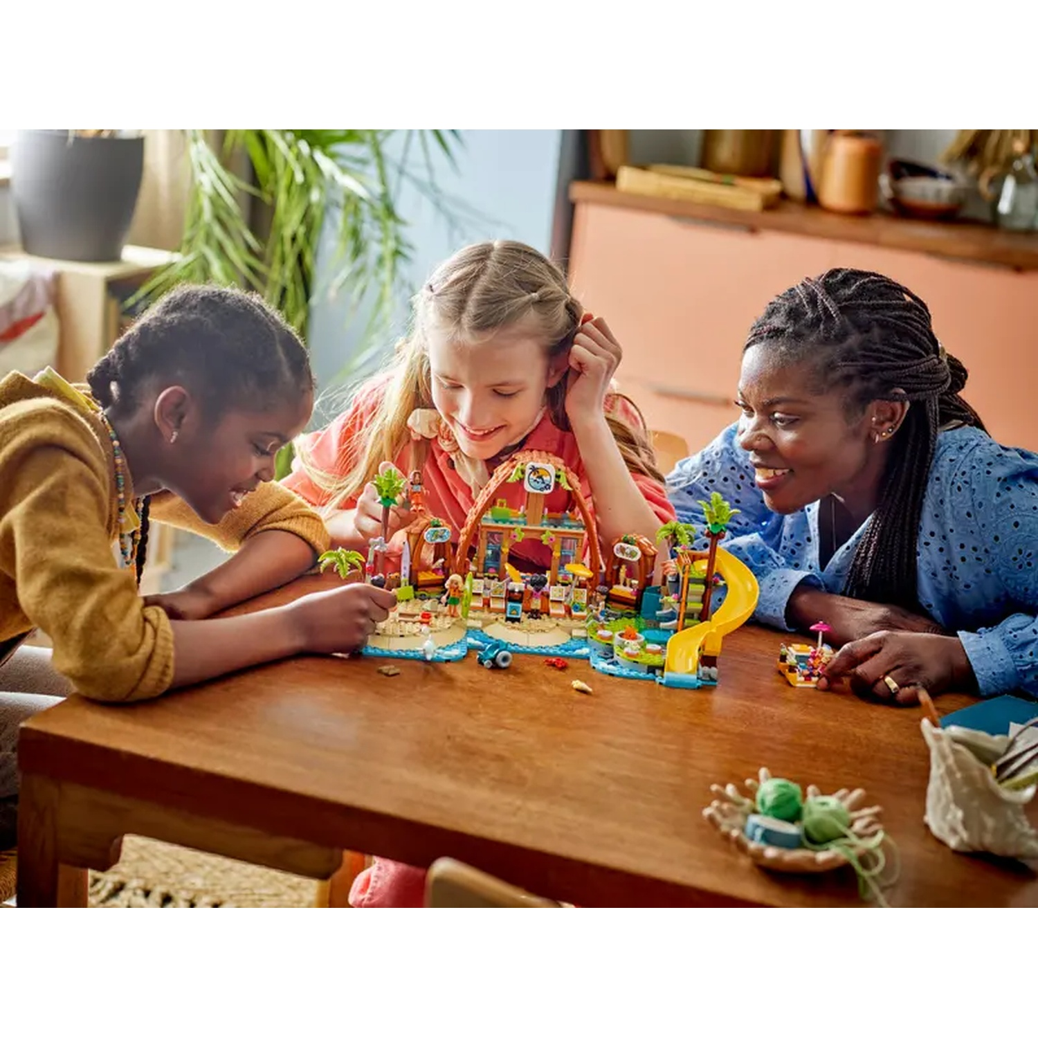 Three girls and a woman play with a Lego set on a wooden table. The set is a beach scene with a slide.