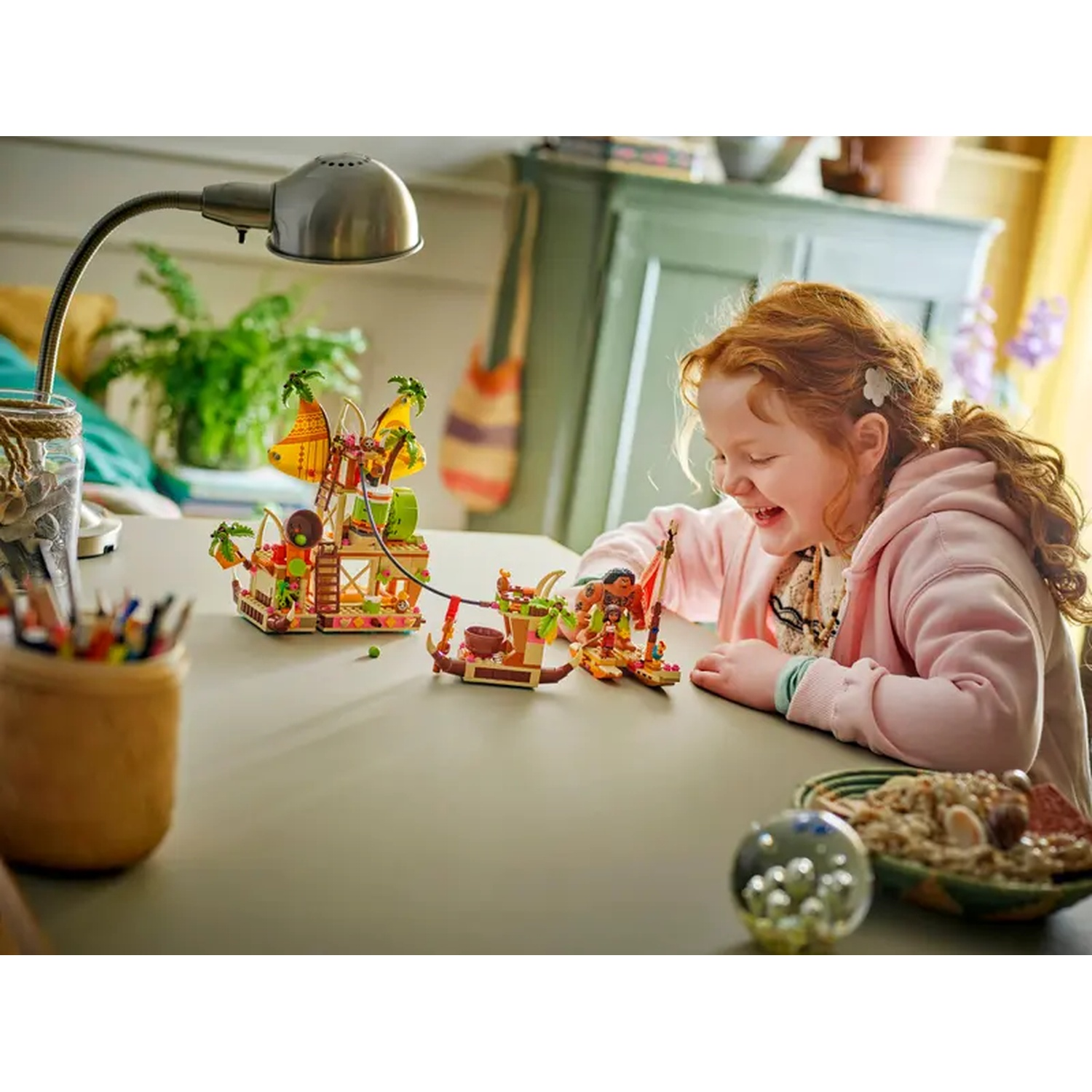 Une fille rit des jeux de jouets sur une table. Une lampe, des plantes et une armoire sont à l'arrière-plan.