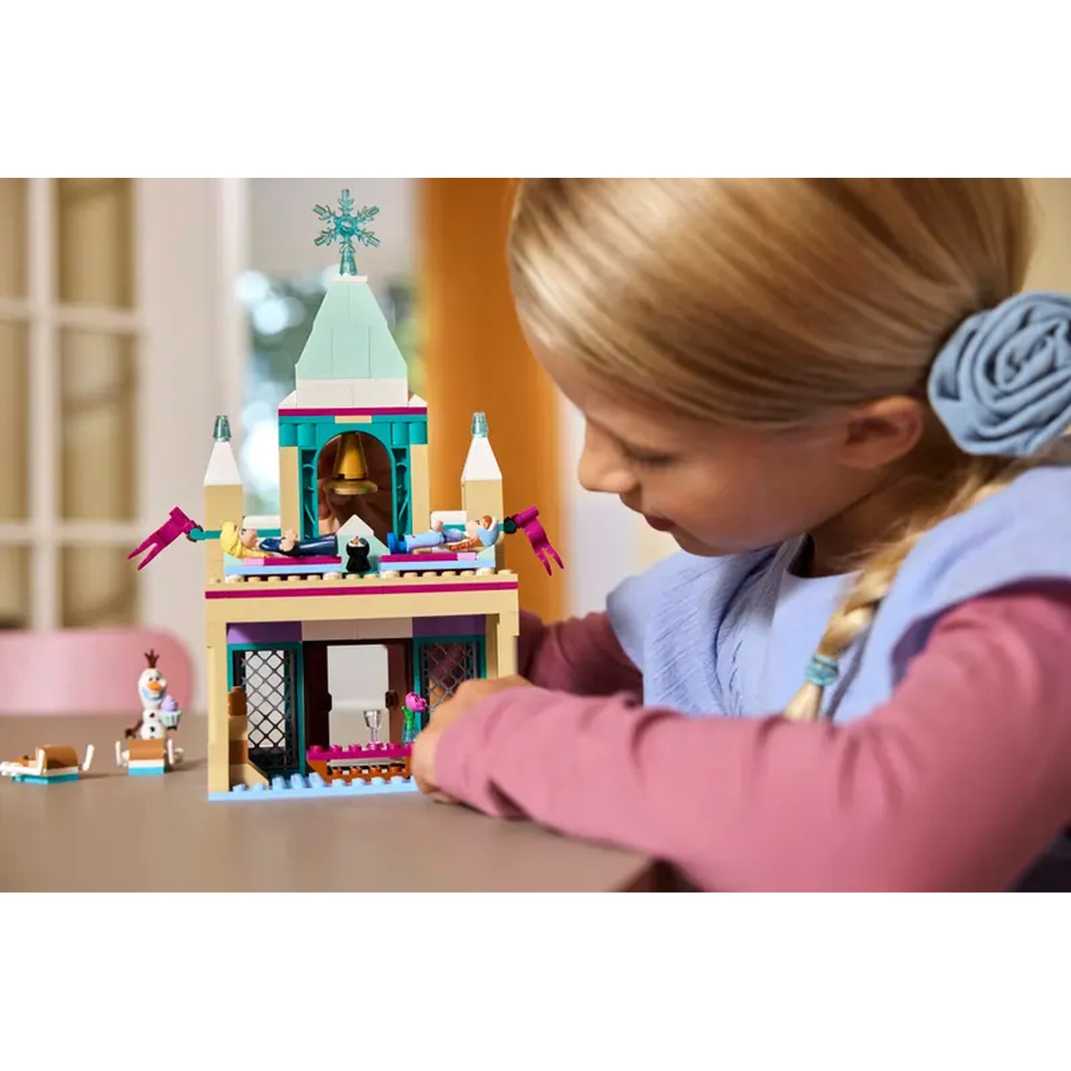 Een meisje bekijkt een Lego-kasteel. Het kasteel is beige, turquoise en roze, met kleine figuurtjes. Olaf en een slee staan ernaast.