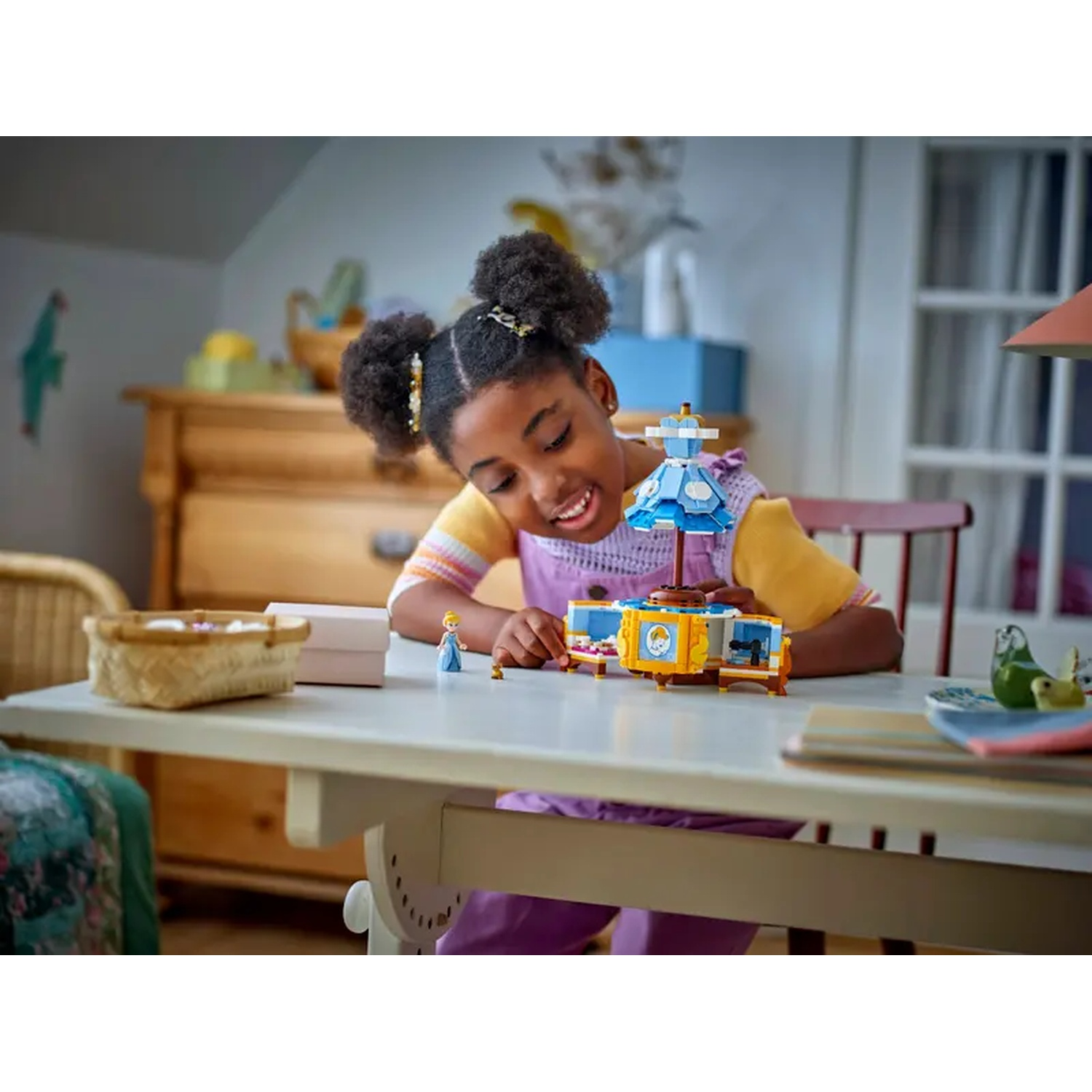 Een meisje glimlacht naar een LEGO-set. De set is een draaimolen. Er staat een dressoir en een mand op tafel.