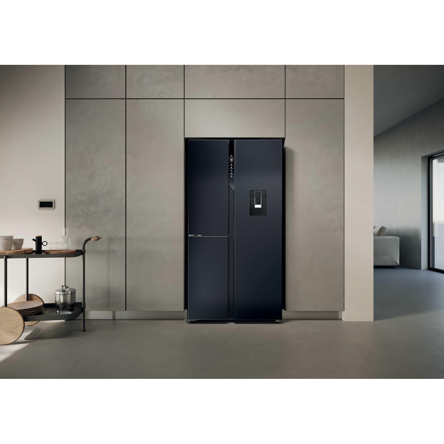 Un frigorifero nero è contro un muro grigio in una cucina moderna. C'è un carrello di servizio a sinistra. Un divano è visibile attraverso una porta.
