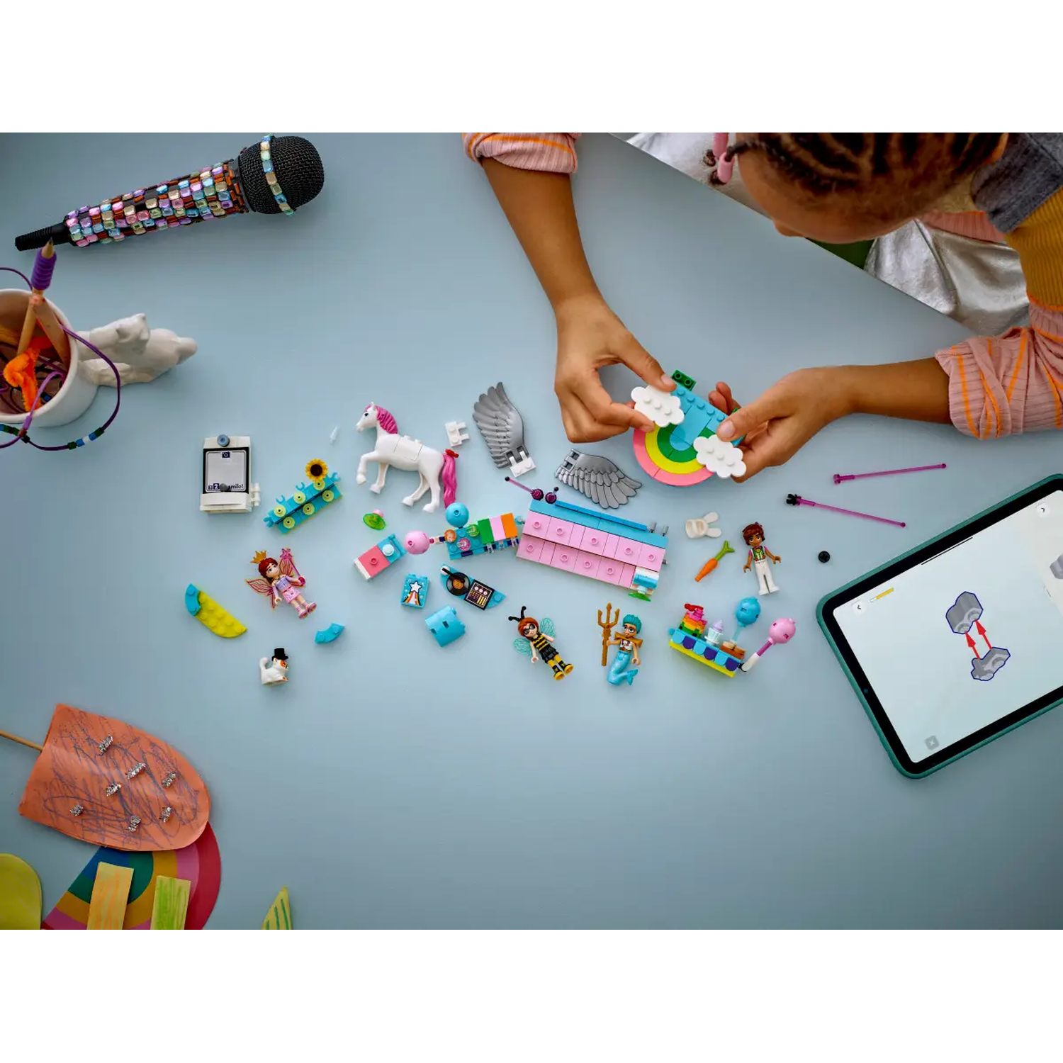A child is assembling a Lego set on a blue surface. Various Lego figures and objects are scattered around. A tablet shows instructions.