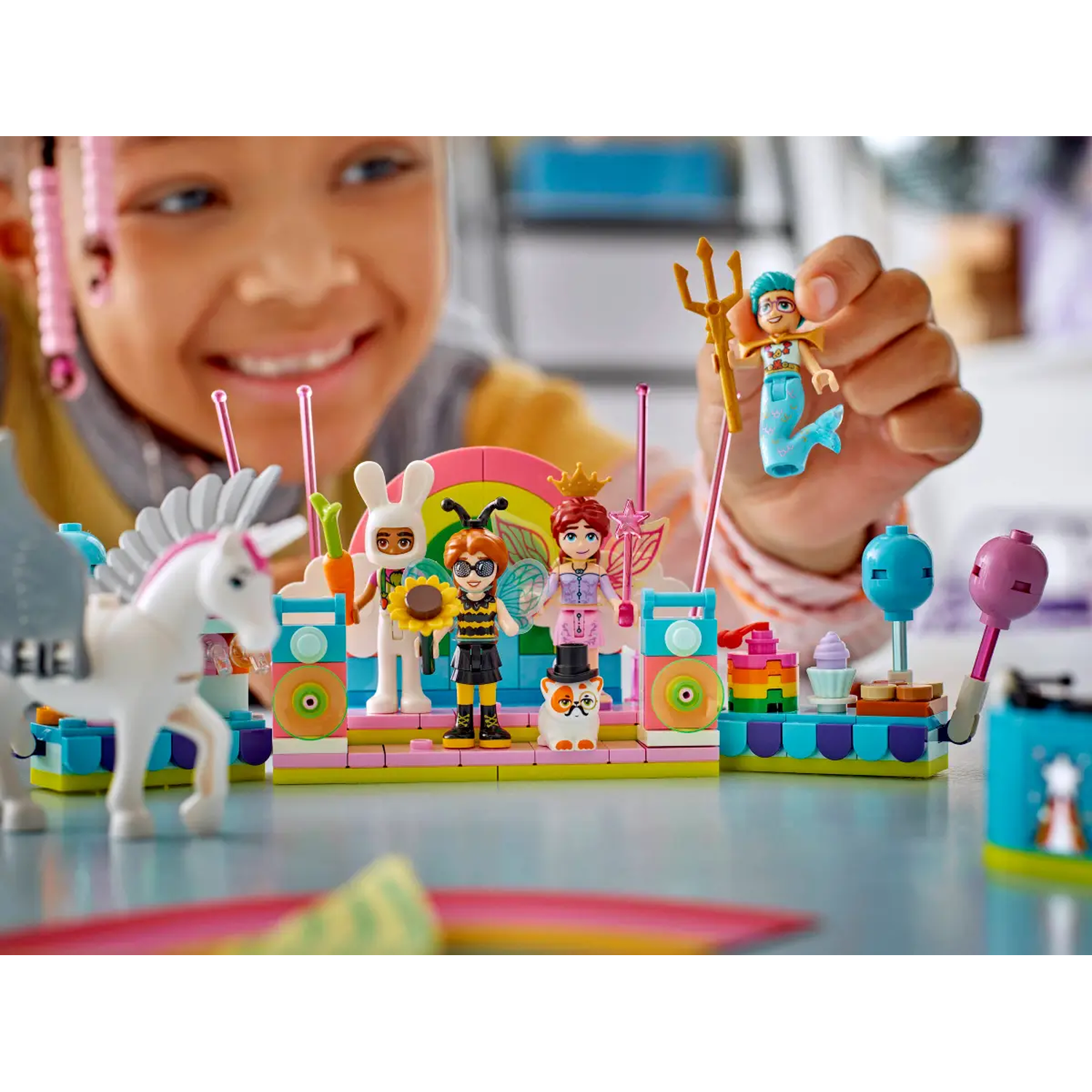 A child holds a toy mermaid. A stage with toy figures of a bunny, bee, and fairy is in front. A white unicorn stands near the stage.
