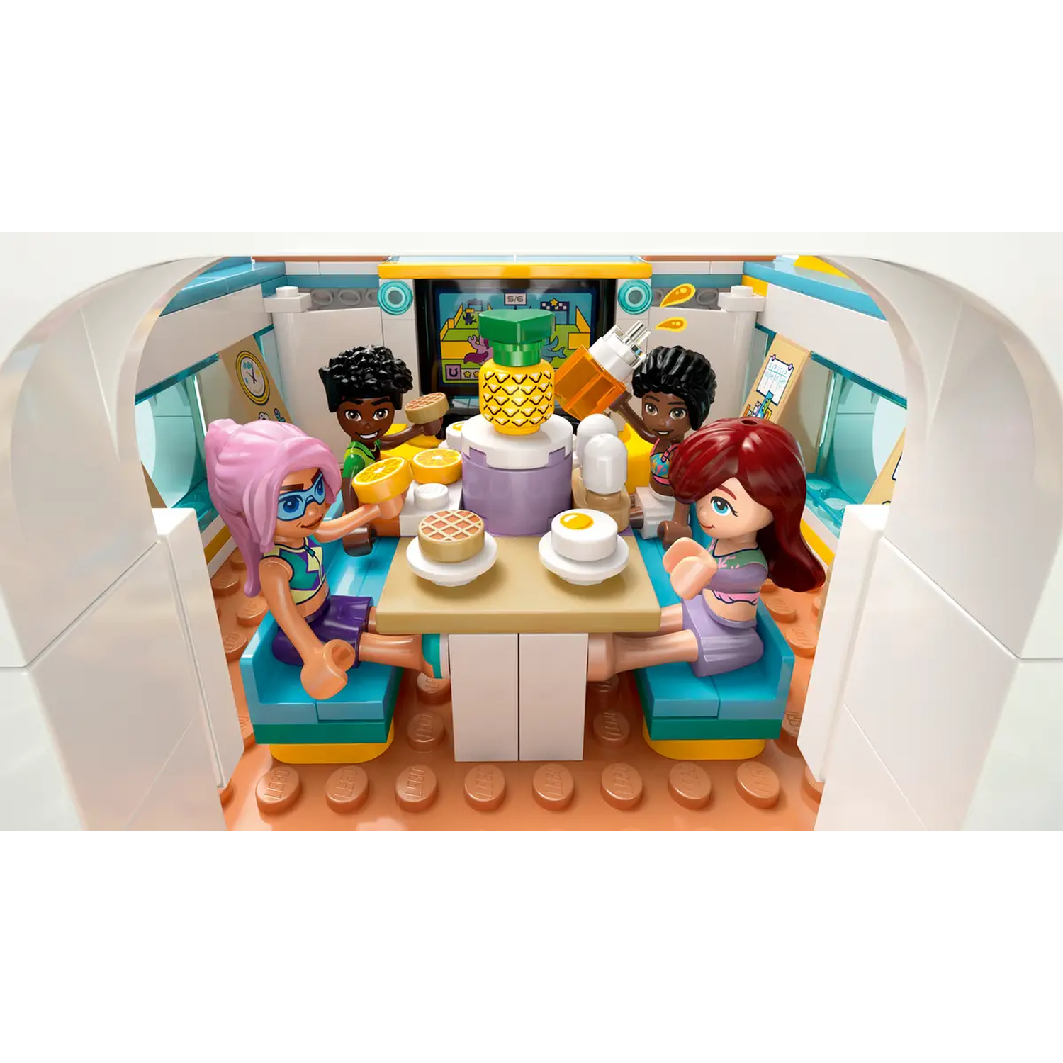 Lego Friends set. Four figures around a table with food, including a pineapple. The scene is inside a white vehicle.