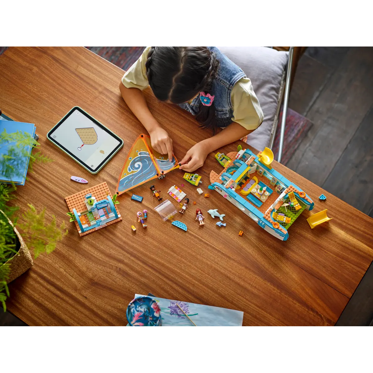 A girl assembles a Lego set on a wooden table. The set includes a boat, house, figures, and instructions on a tablet.