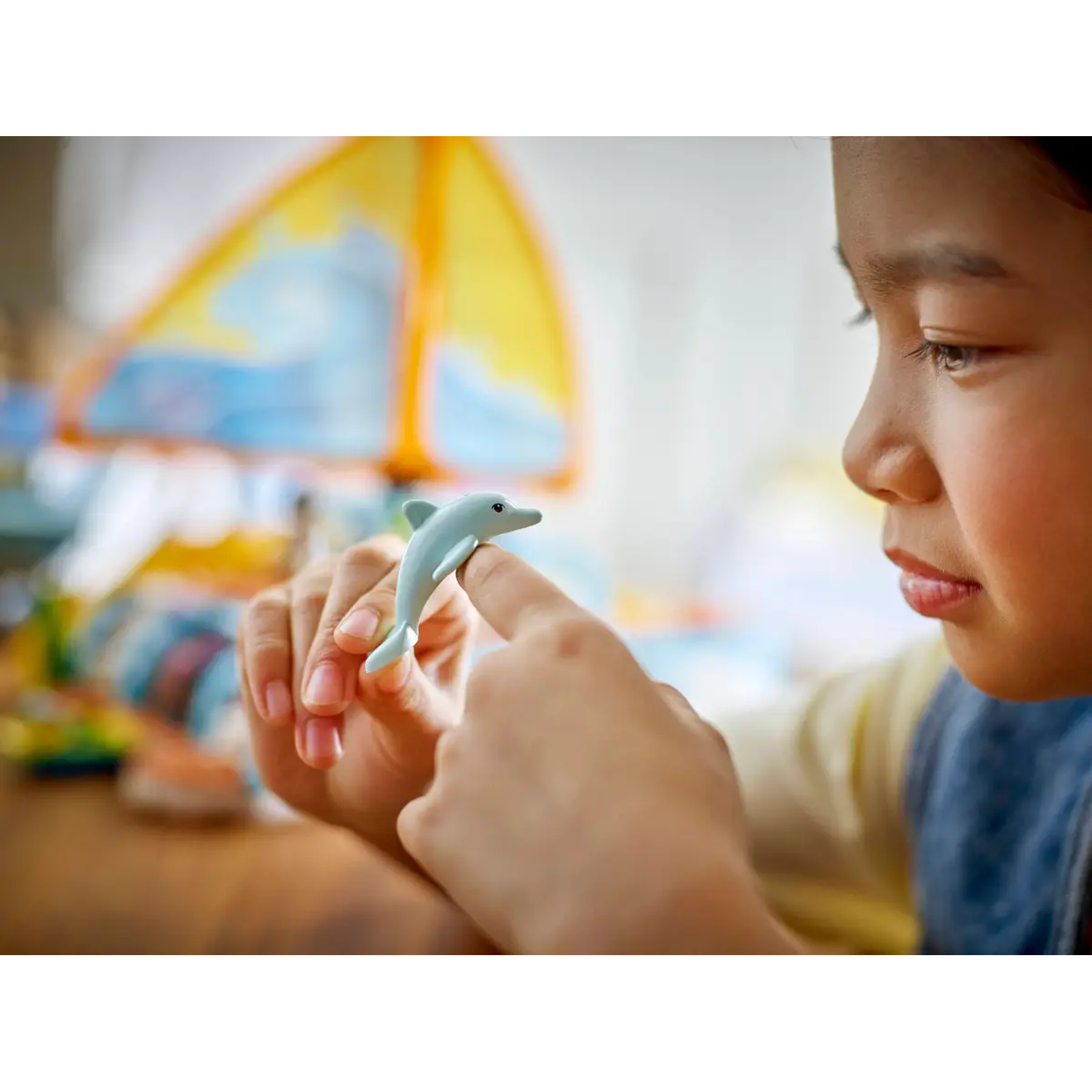 A young girl holds a small blue dolphin figurine, pointing at it. The background has a tent and a blurry setting.
