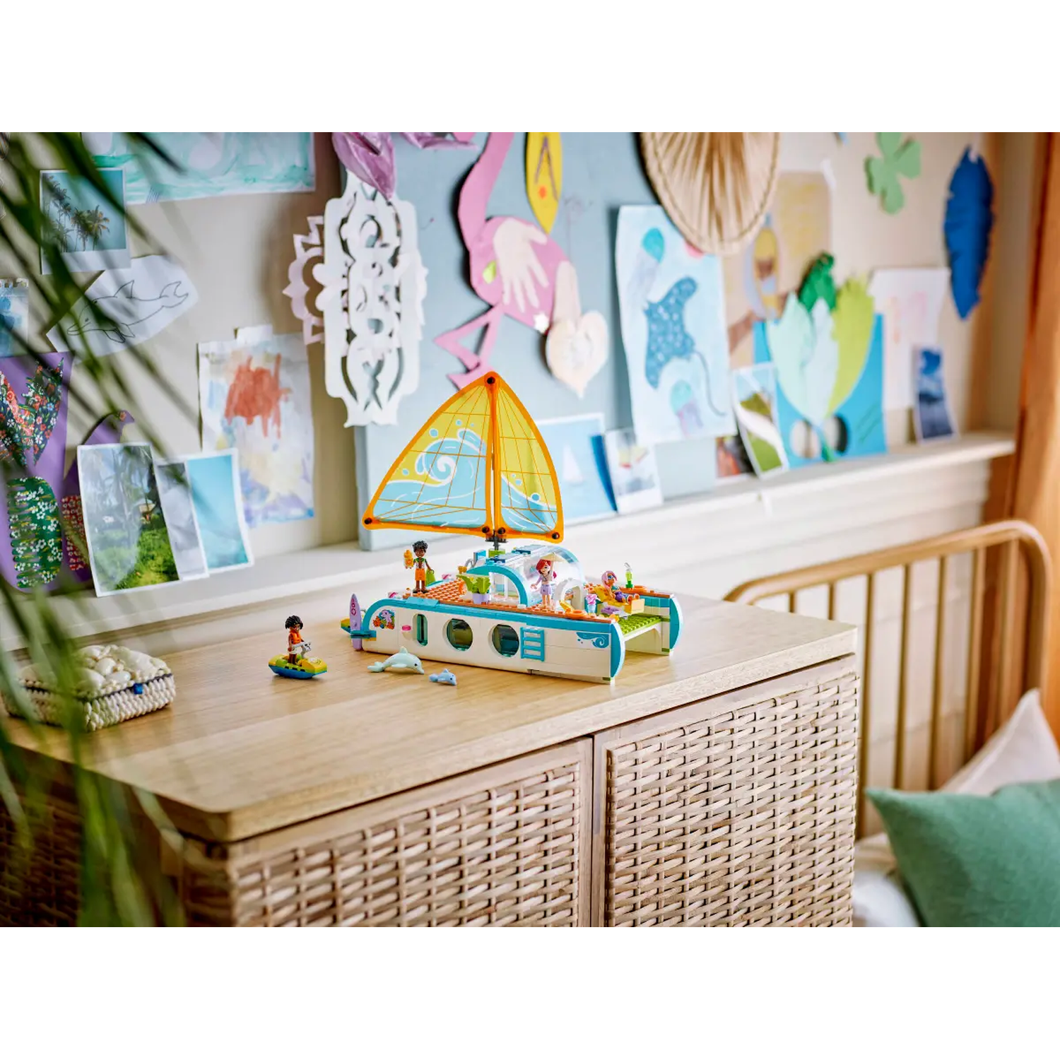 A Lego sailboat on a wooden table. The background shows drawings and a wooden headboard. A green pillow is on the right.