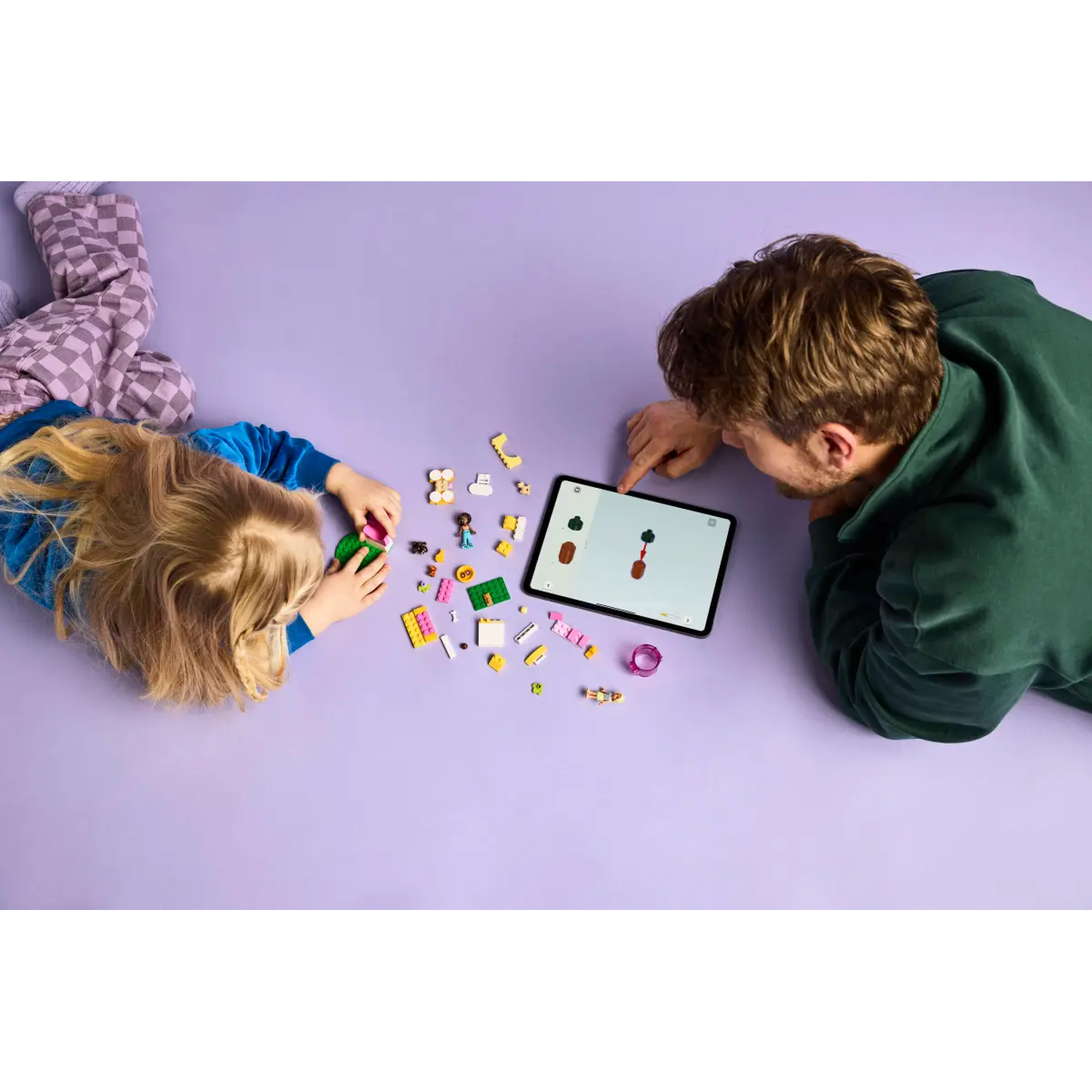 A child and an adult are assembling Lego toys on a purple surface, looking at a tablet. Toys and Lego bricks are scattered around them.